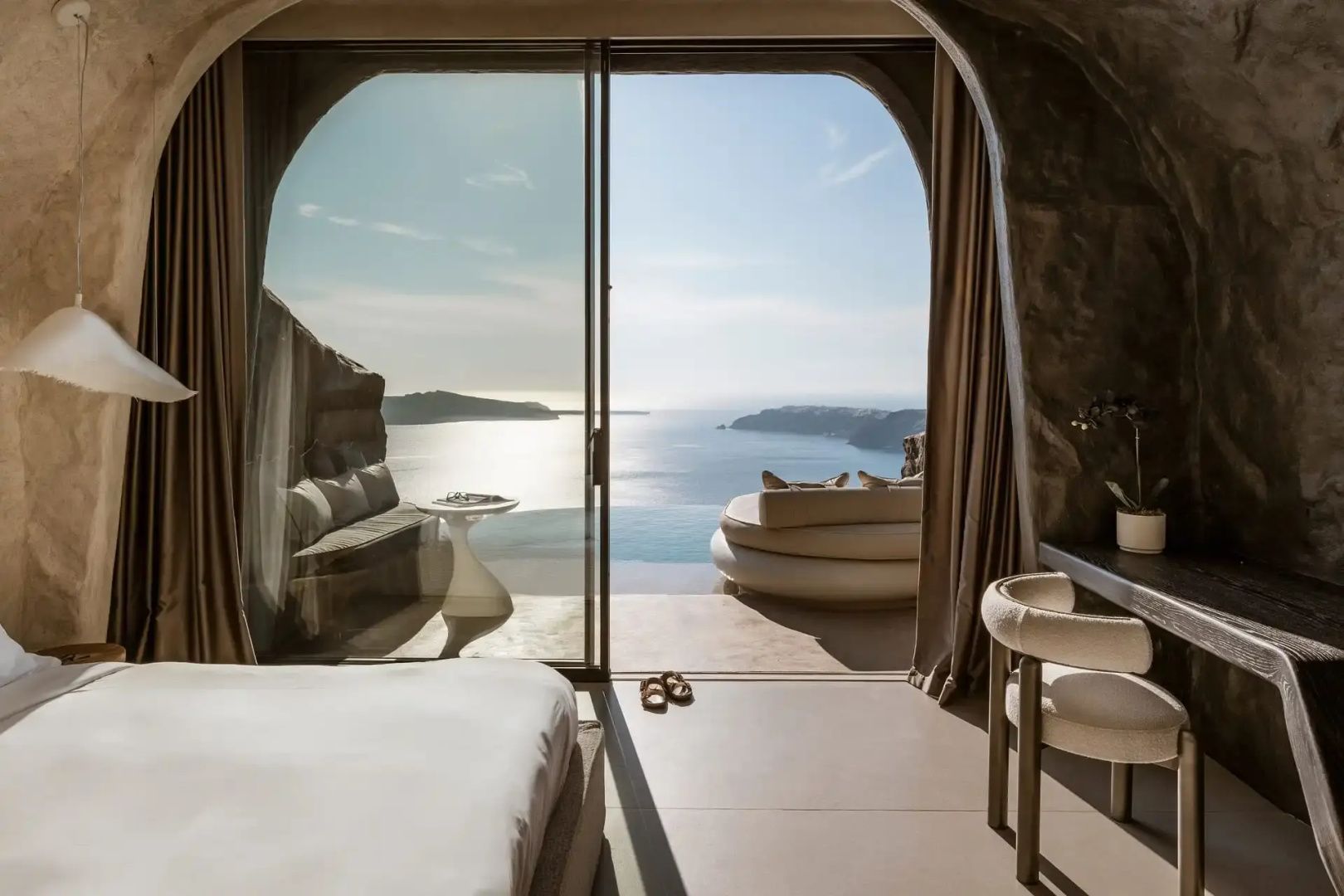 Blick durch das große Glasfenster der Suite auf die Terrasse mit Liegebett und das Meer.