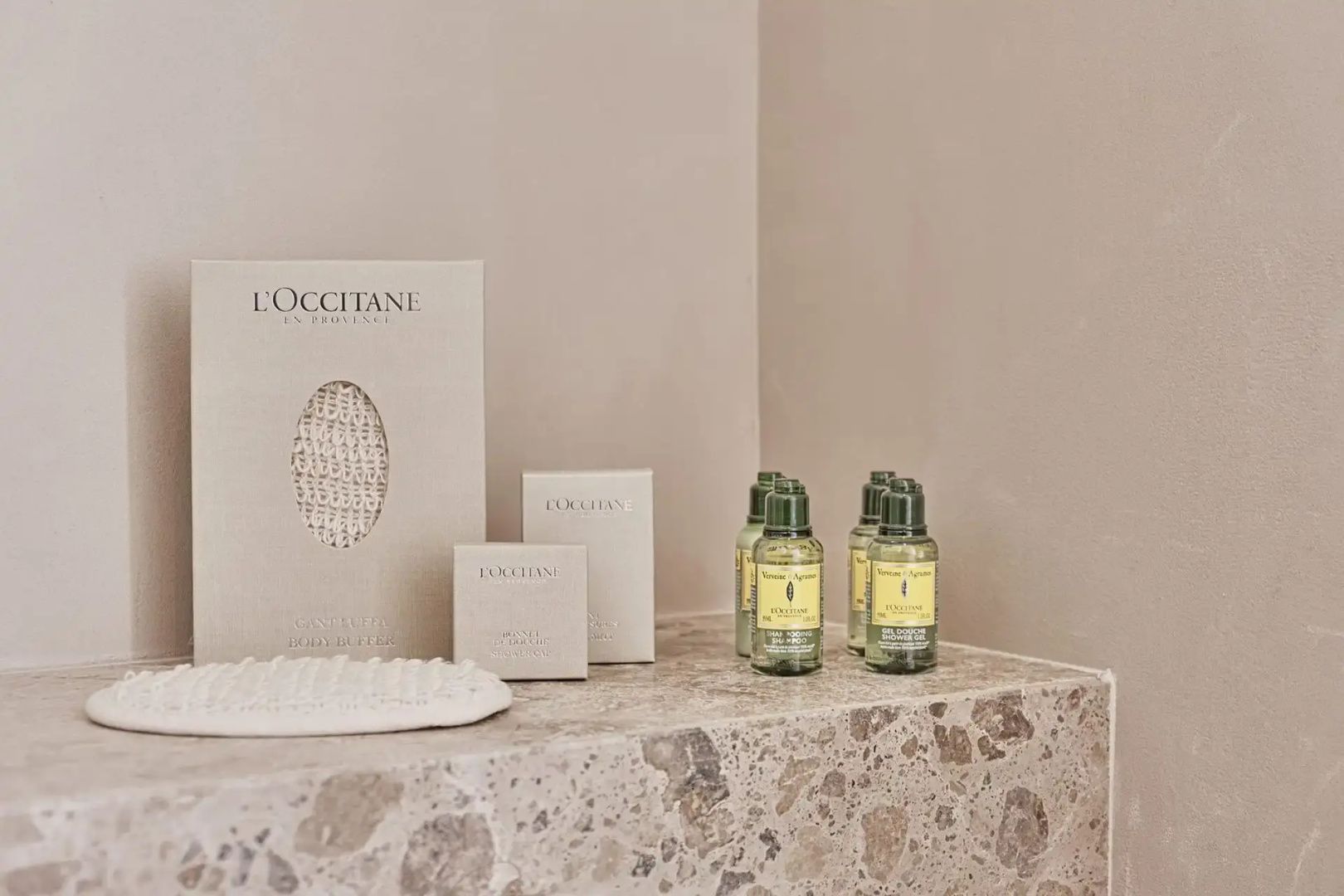 Pflegeprodukte von L'Occitane und ein Pflegeschwamm auf einer hellen Steinablage.