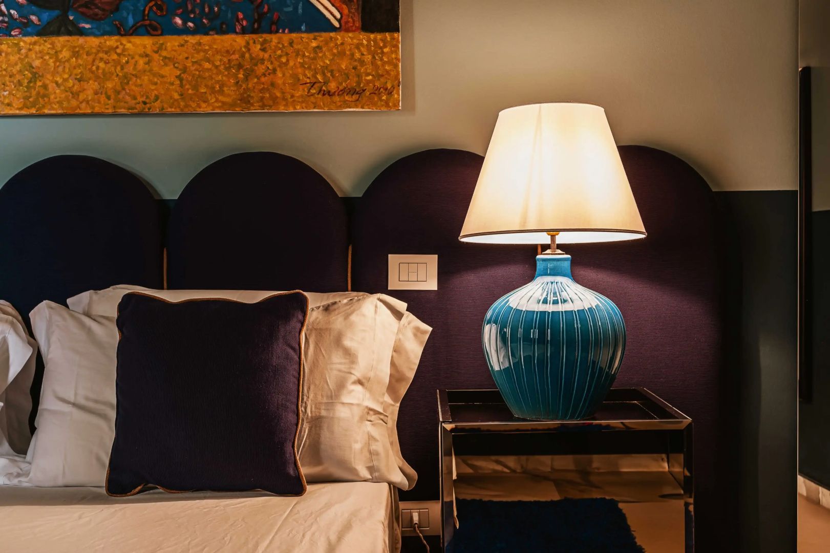 Nachttisch mit leuchtender blauer Keramiktischlampe neben einem Bett mit blauem Kopfteil.