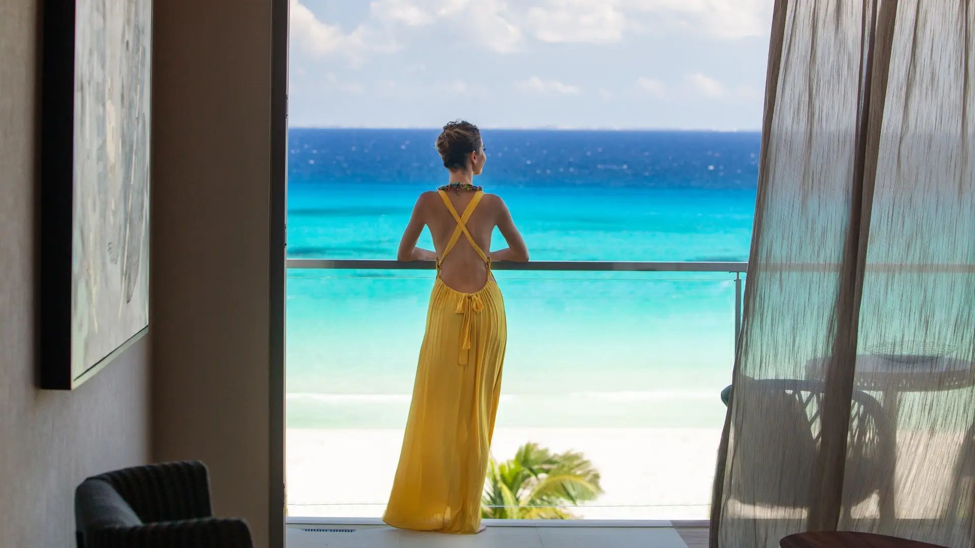 Frau im gelben Kleid blickt von einem Balkon auf das türkisfarbene Meer und den Strand.