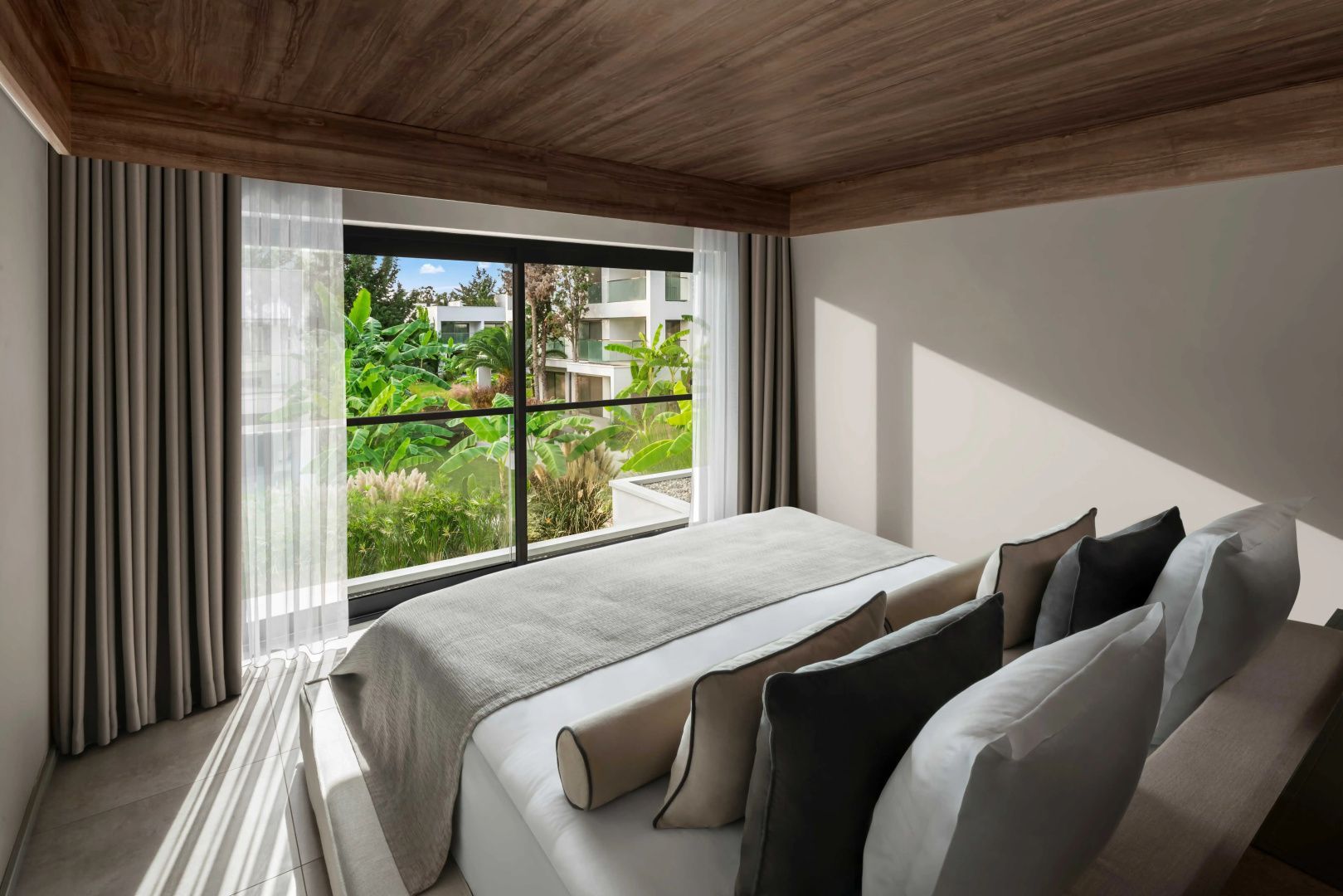 Doppelbett mit hellgrauer Bettwäsche vor einer Fensterfront mit Blick ins Grüne.