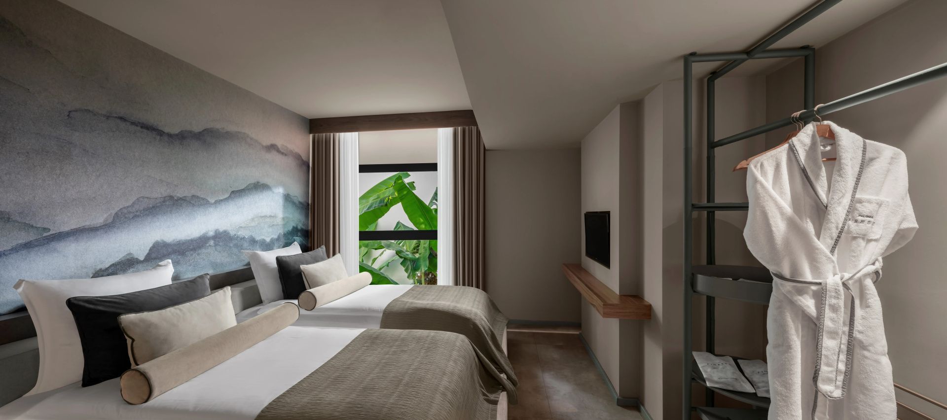 Hotelzimmer mit zwei Betten, beigen Tagesdecken, einem Wandfernseher und Fenster mit Blick auf Grünpflanzen.
