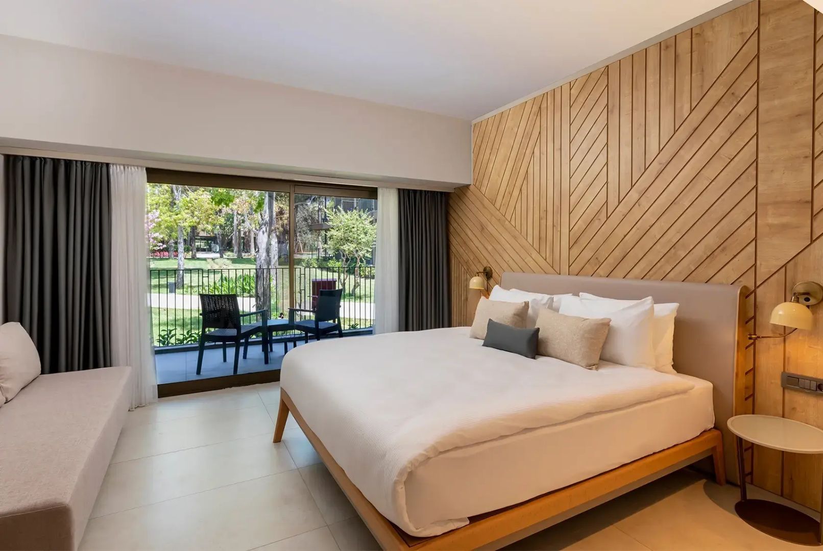 Doppelbett mit hellen Kissen und eine Glasfront mit Blick auf einen Balkon im Hotelzimmer.