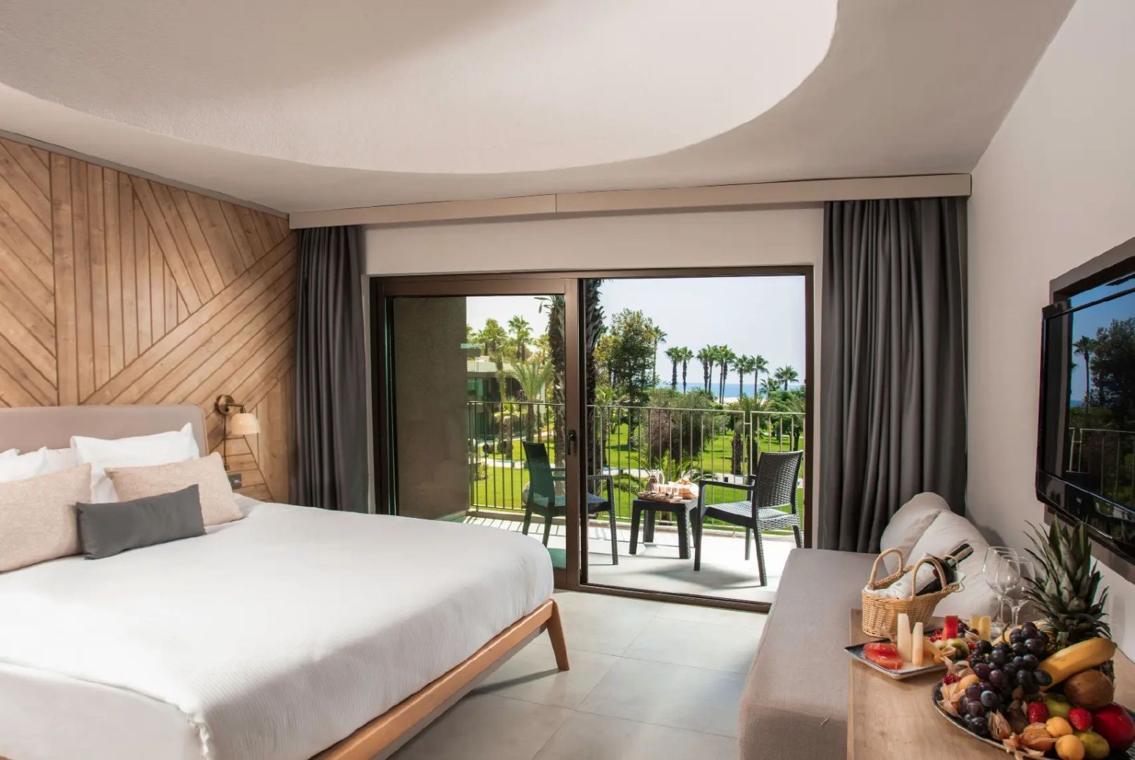 Modernes Hotelzimmer mit Doppelbett, Sofa und Balkon mit Blick auf Palmen und Gartenanlagen.