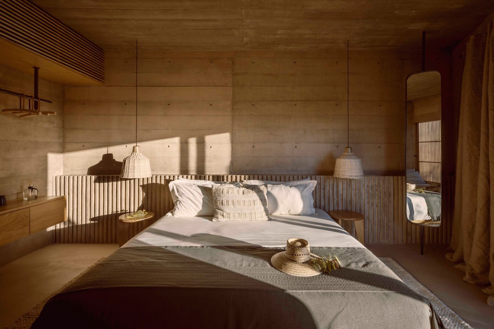 Schlafzimmer mit Doppelbett, beigefarbenen Betonwänden und zwei geflochtenen Hängelampen.