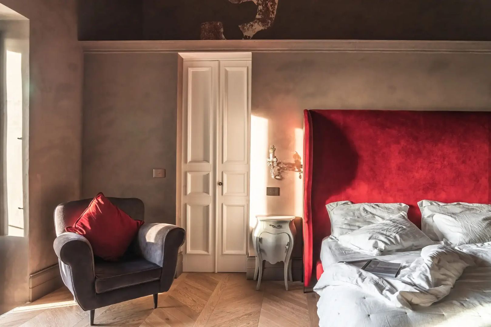 Hotelzimmer mit rotem Samtkopfteil am Bett, Sessel, Nachttisch und Holzparkett.