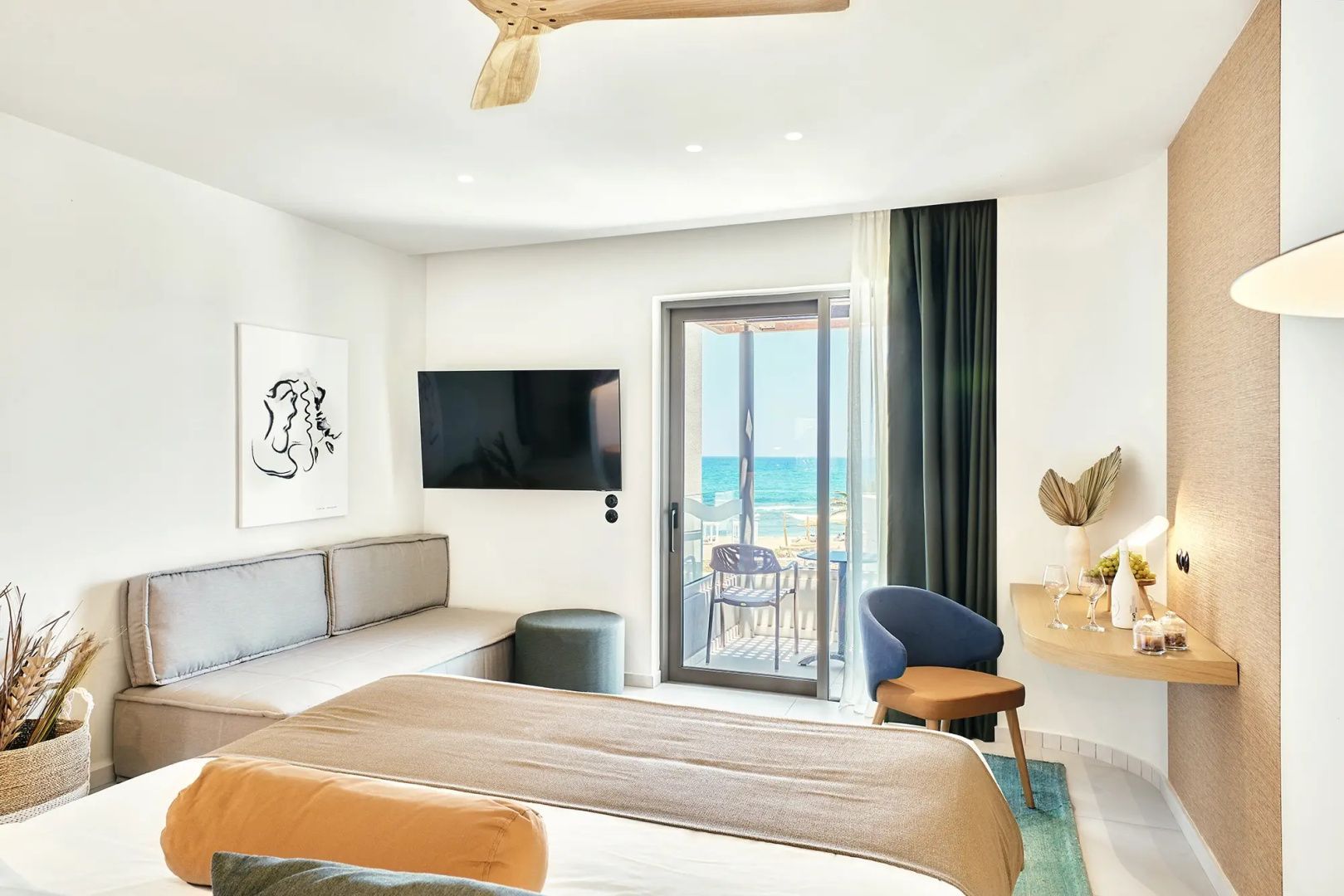 Blick durch die Glasbalkontür eines hellen Hotelzimmers auf den Strand und das blaue Meer.