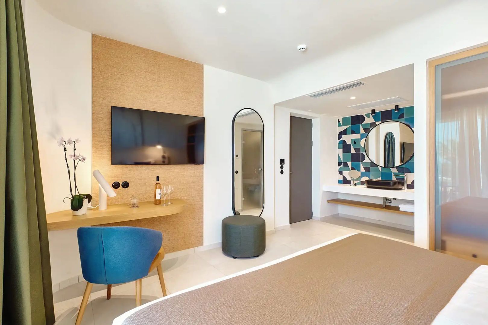 Wohnbereich eines Hotelzimmers mit Fernseher, Schreibtisch, Spiegel und offenem Waschbereich.