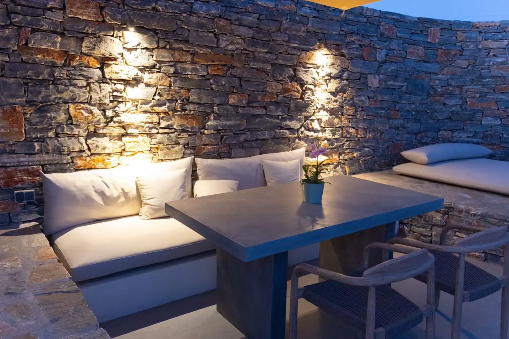 Überdachte Terrasse mit Steinwand, beleuchtetem Esstisch und bequemer Sitzbank.