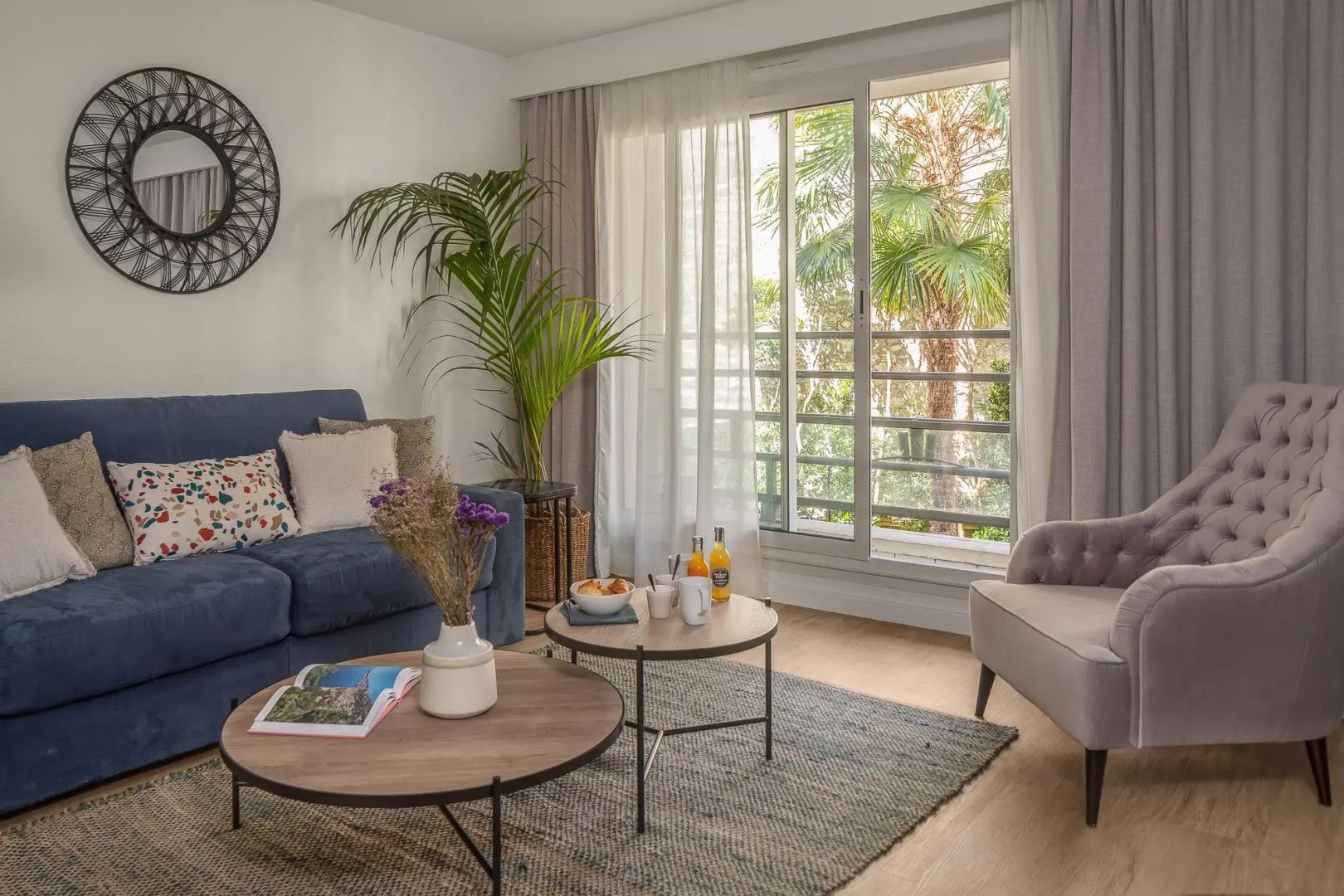 Modernes Wohnzimmer mit blauem Sofa, zwei Couchtischen und Pflanzen am Fenster.