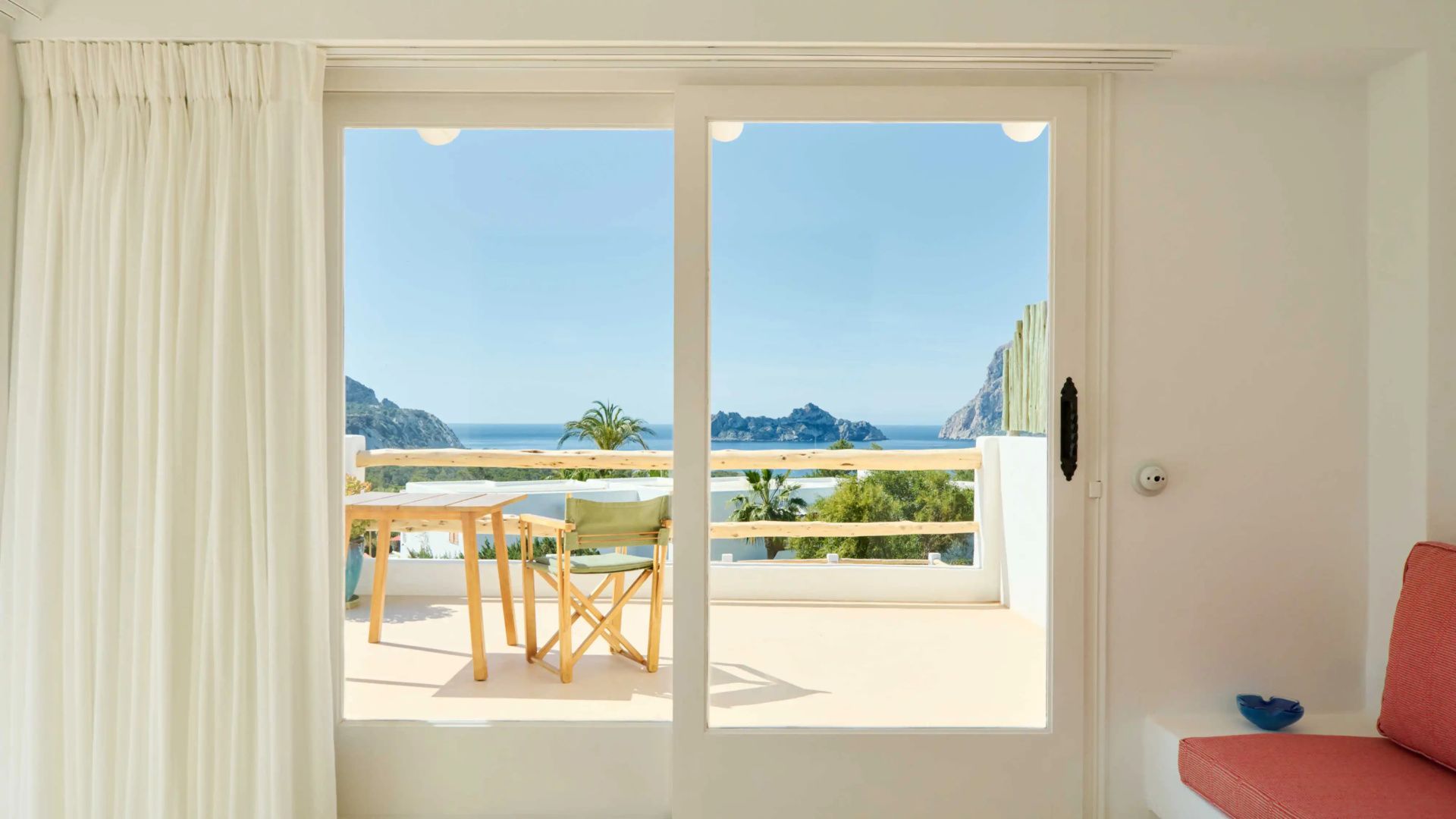 Blick durch eine Schiebetür auf eine weiße Terrasse mit Holztisch und dem Meer im Hintergrund.