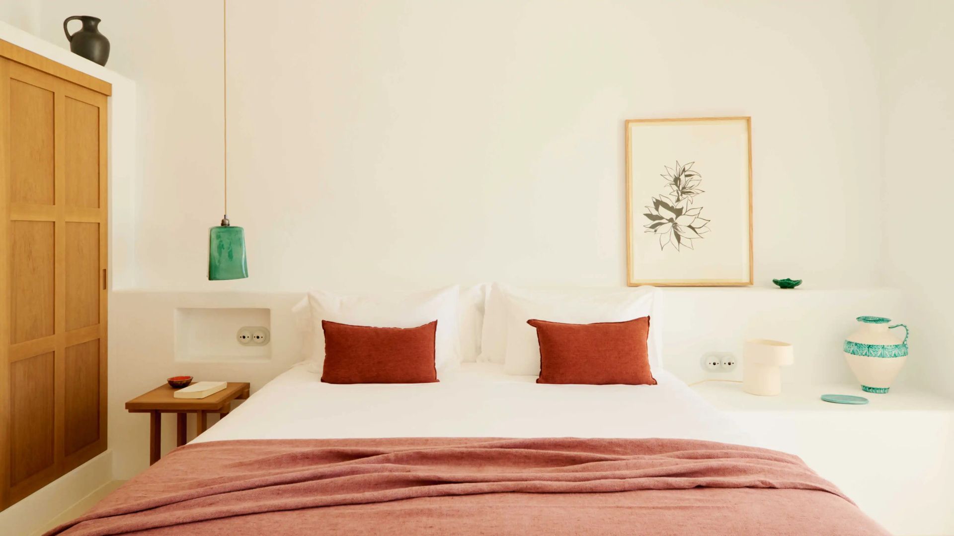 Doppelbett mit weißen Kissen, roter Tagesdecke und hellem Wandbild im Hotelzimmer.