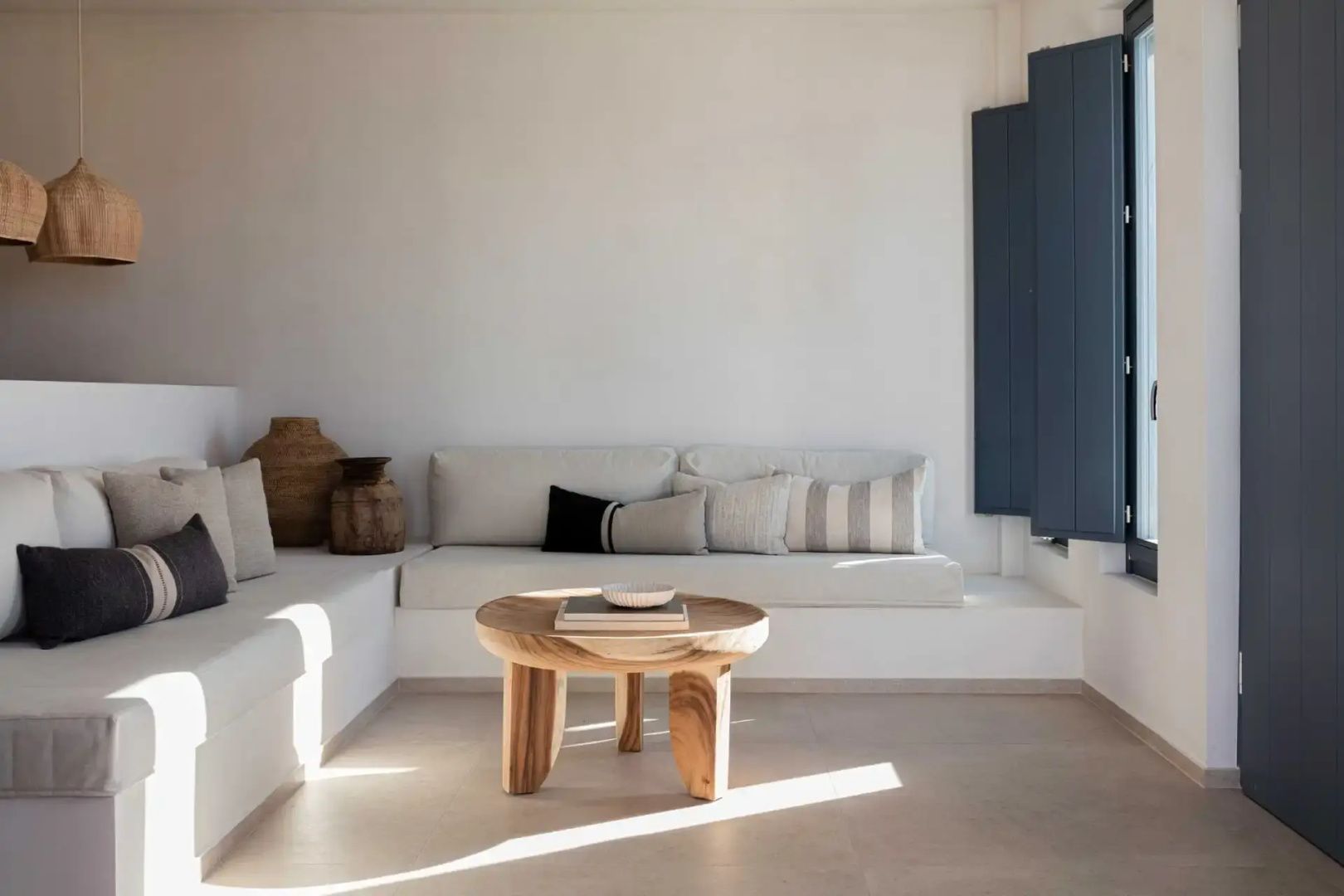 Hell eingerichteter Wohnbereich mit weißem Einbau-Sofa, Kissen und einem runden Holztisch.