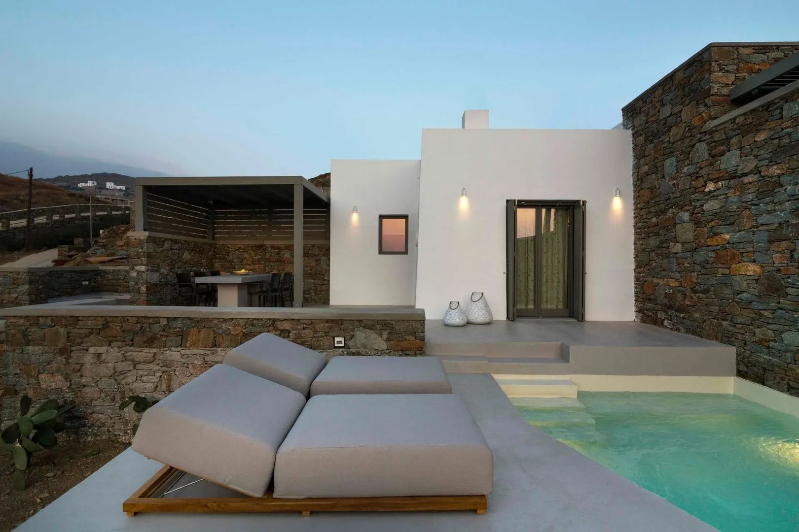 Moderne weiße Villa mit Natursteinmauer, Sonnenliegen und Swimmingpool am Abend.