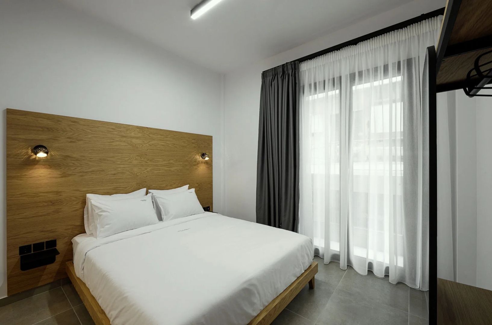 Doppelbett mit weißer Bettwäsche, Holzrückwand und bodentiefen Fenstern mit Vorhängen.
