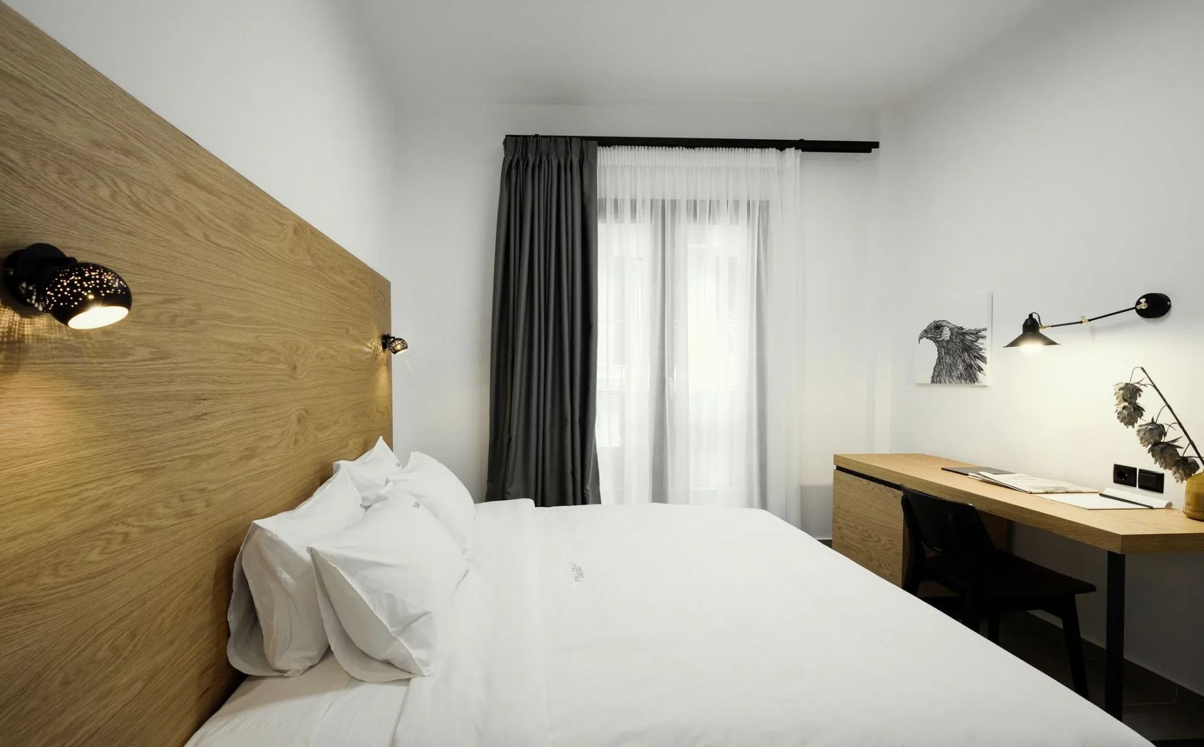 Doppelbett mit weißer Bettwäsche, hölzernem Kopfteil und grauen Vorhängen am Fenster.