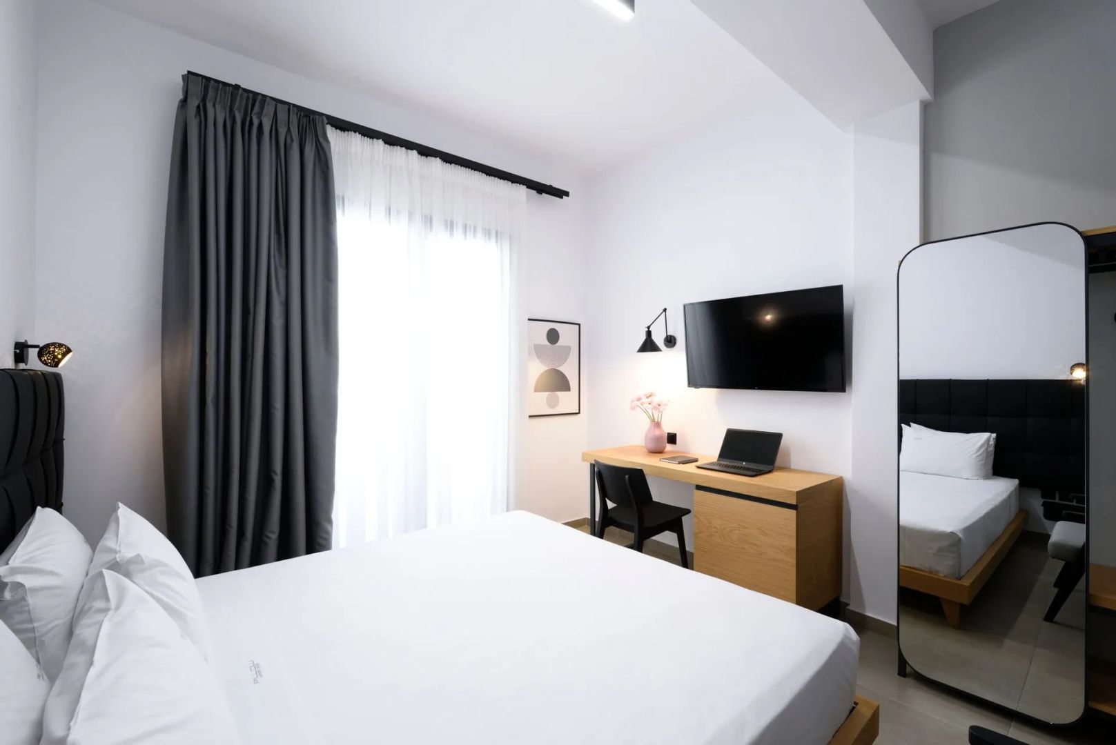 Helles Schlafzimmer mit Holzschreibtisch, Wandfernseher und großem Fenster hinter Vorhängen.