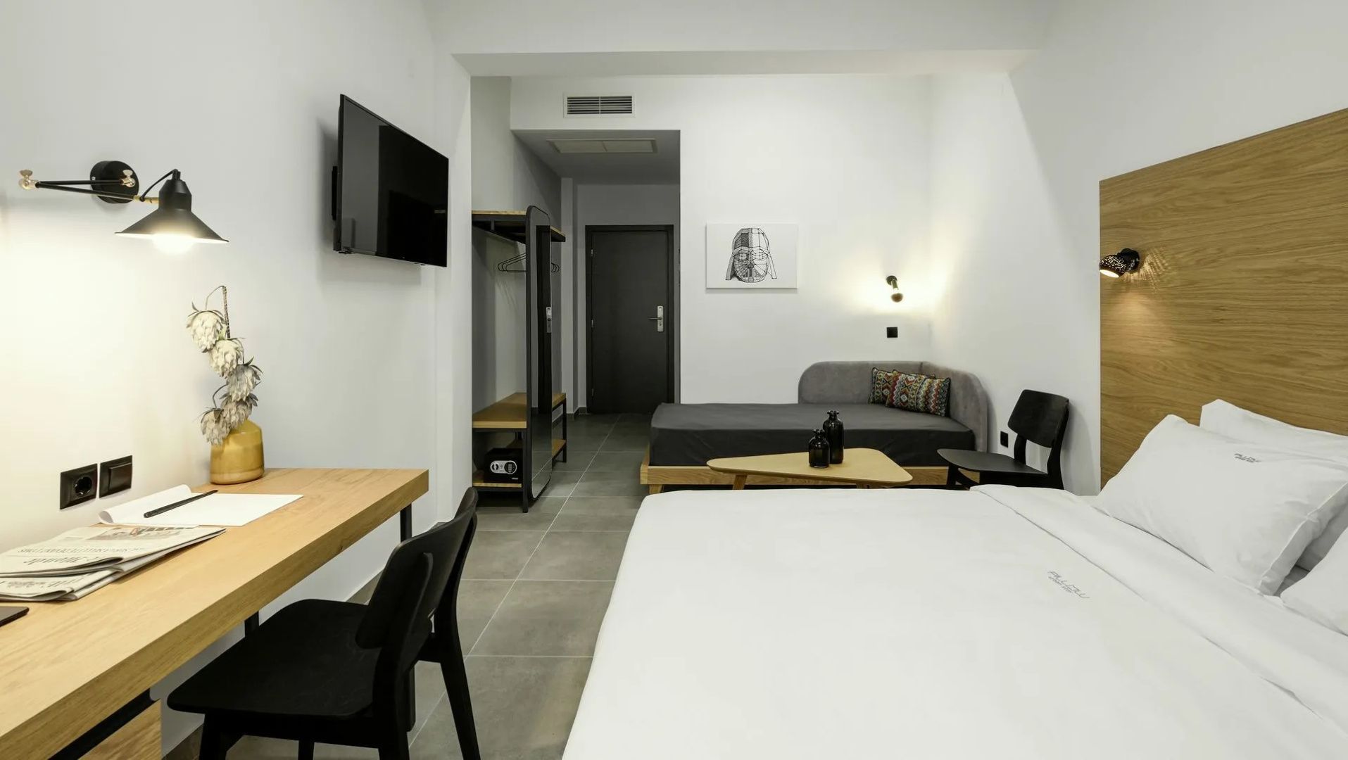 Blick durch das Hotelzimmer auf einen Wandfernseher, ein graues Daybed und die Eingangstür.