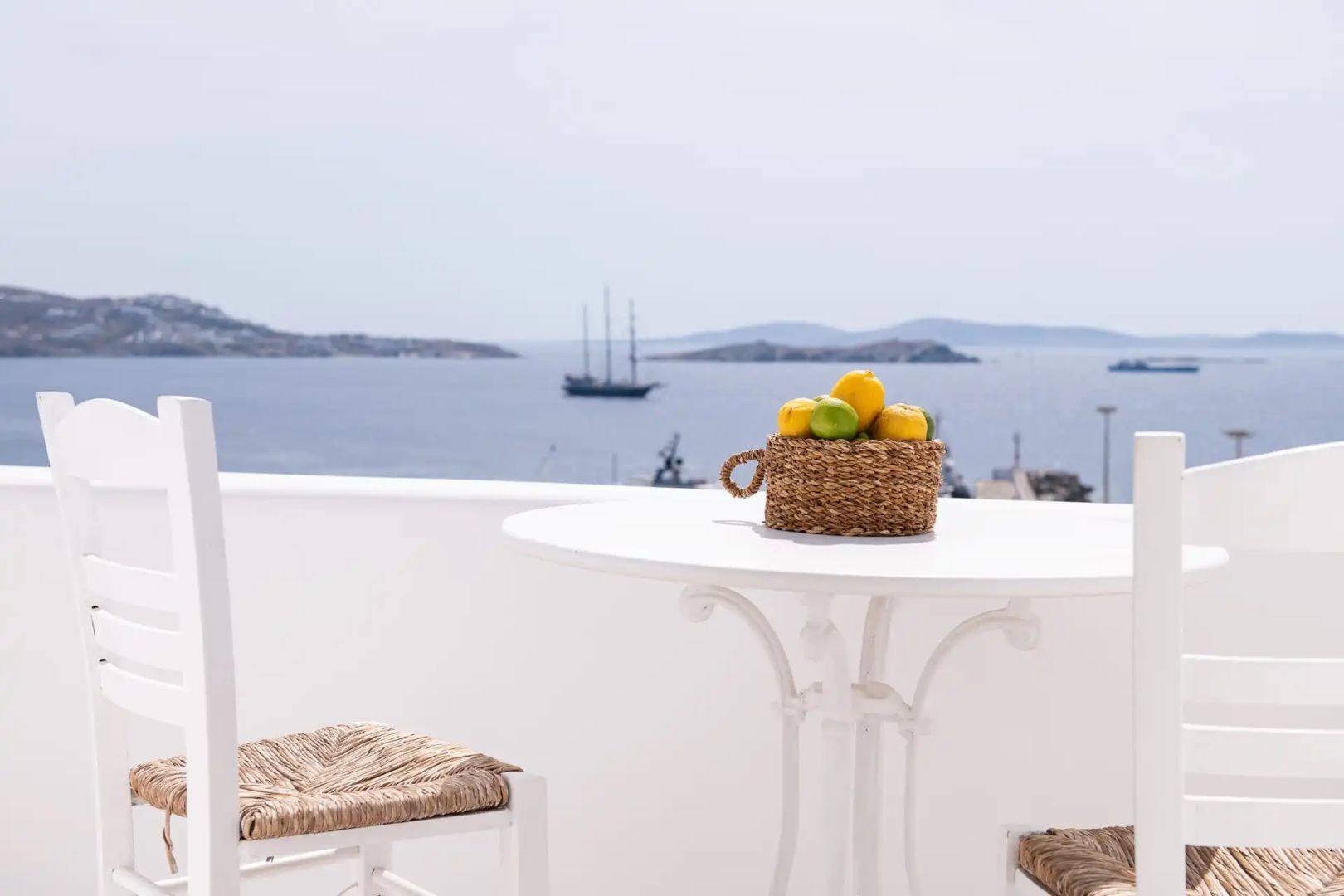 Balkon mit weißem Tisch, Obstkorb und Stühlen vor Meerblick mit Schiffen.