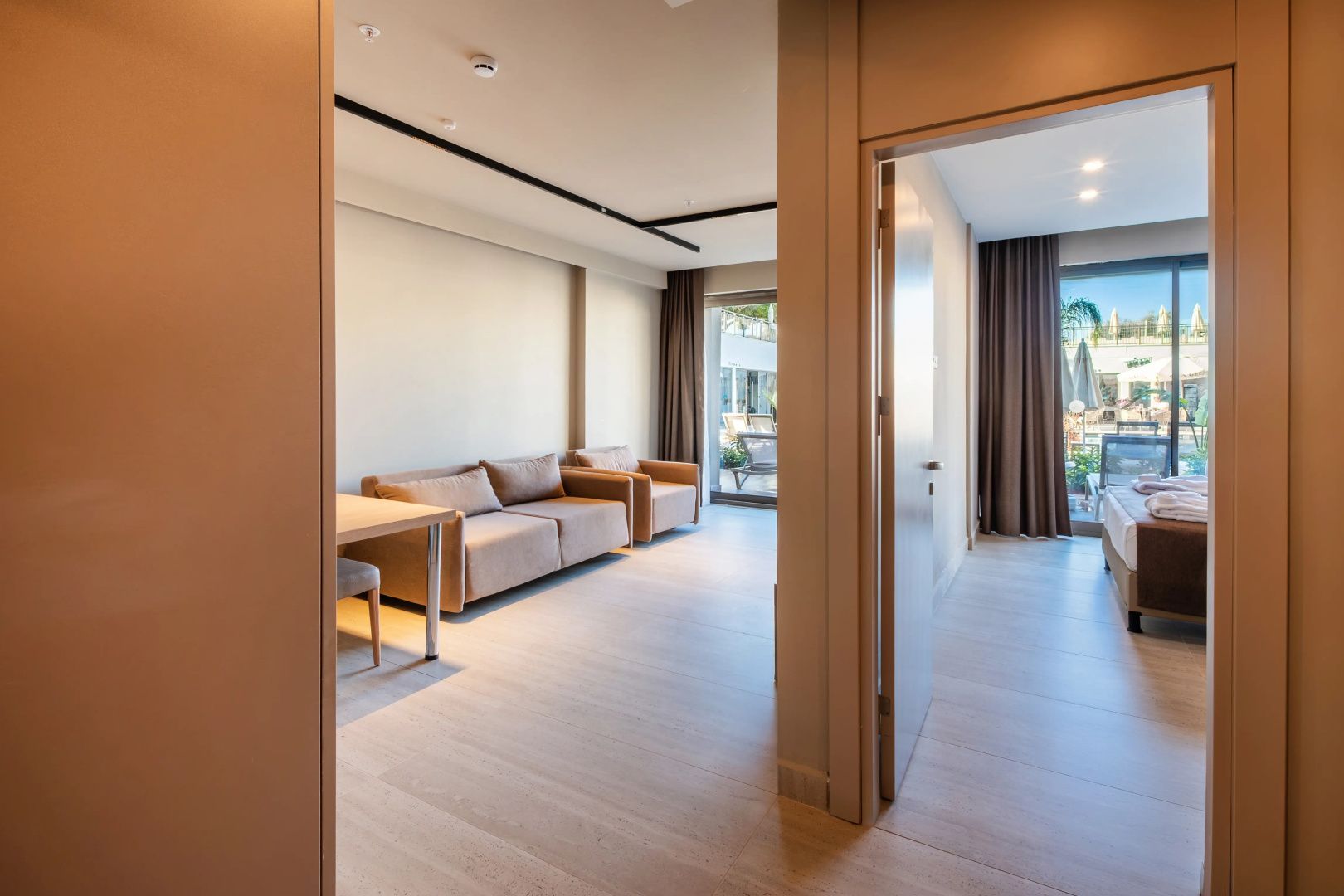 Wohnbereich einer Suite mit beigen Sofas, Holztisch und Zugang zur Terrasse durch eine Balkontür.