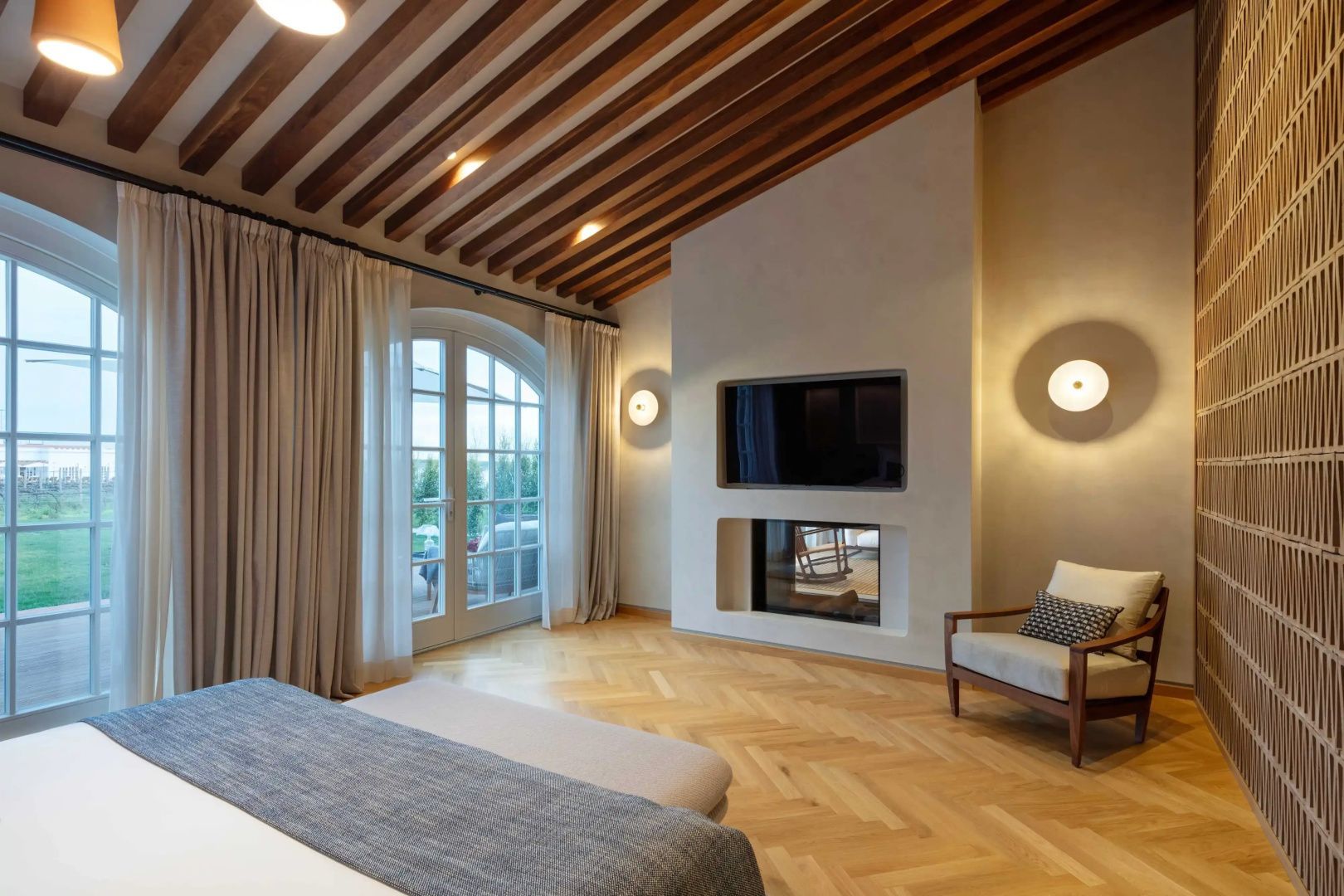 Schlafbereich mit Fernseher an der Wand, Kamin, Sessel und großer Glastür zur Terrasse.