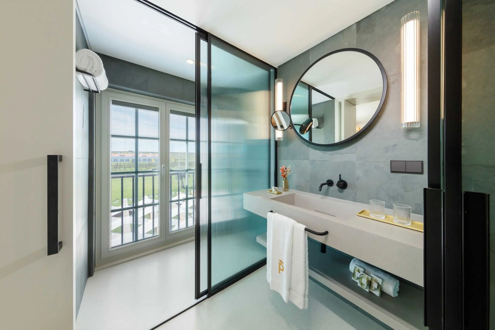 Modernes Badezimmer mit Waschtisch, rundem Wandspiegel und Abtrennung zur Dusche aus Glas.