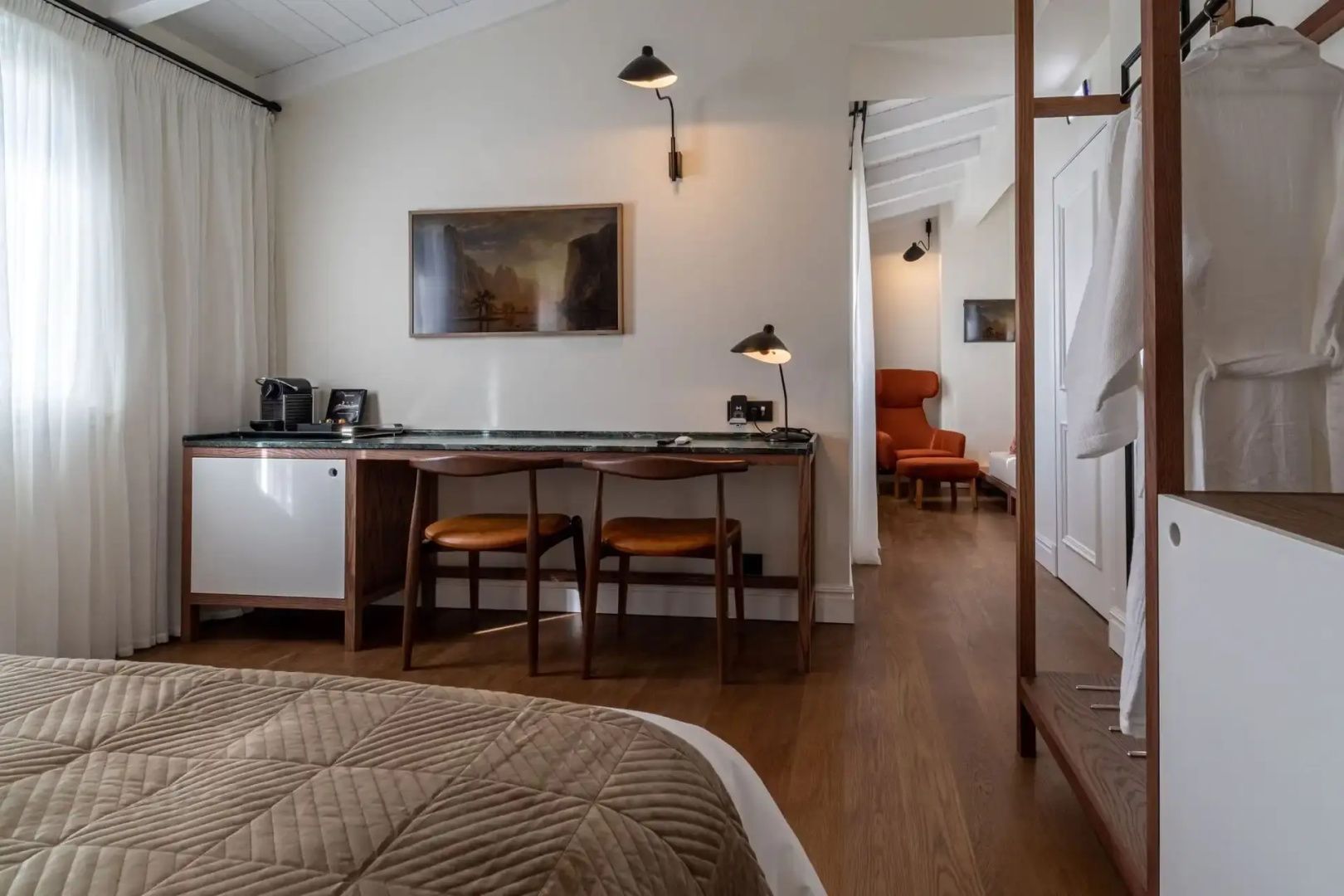 Schreibtisch aus Holz mit zwei Stühlen, Tischlampe und Bild an der hellen Wand.