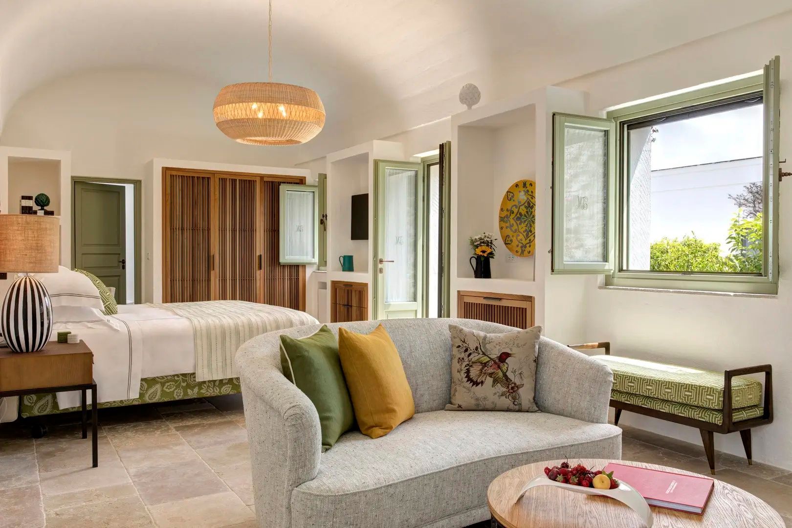 Sitzbereich einer Suite mit grauem Sofa, bunten Kissen, Holzschrank und Blick zum Bett.