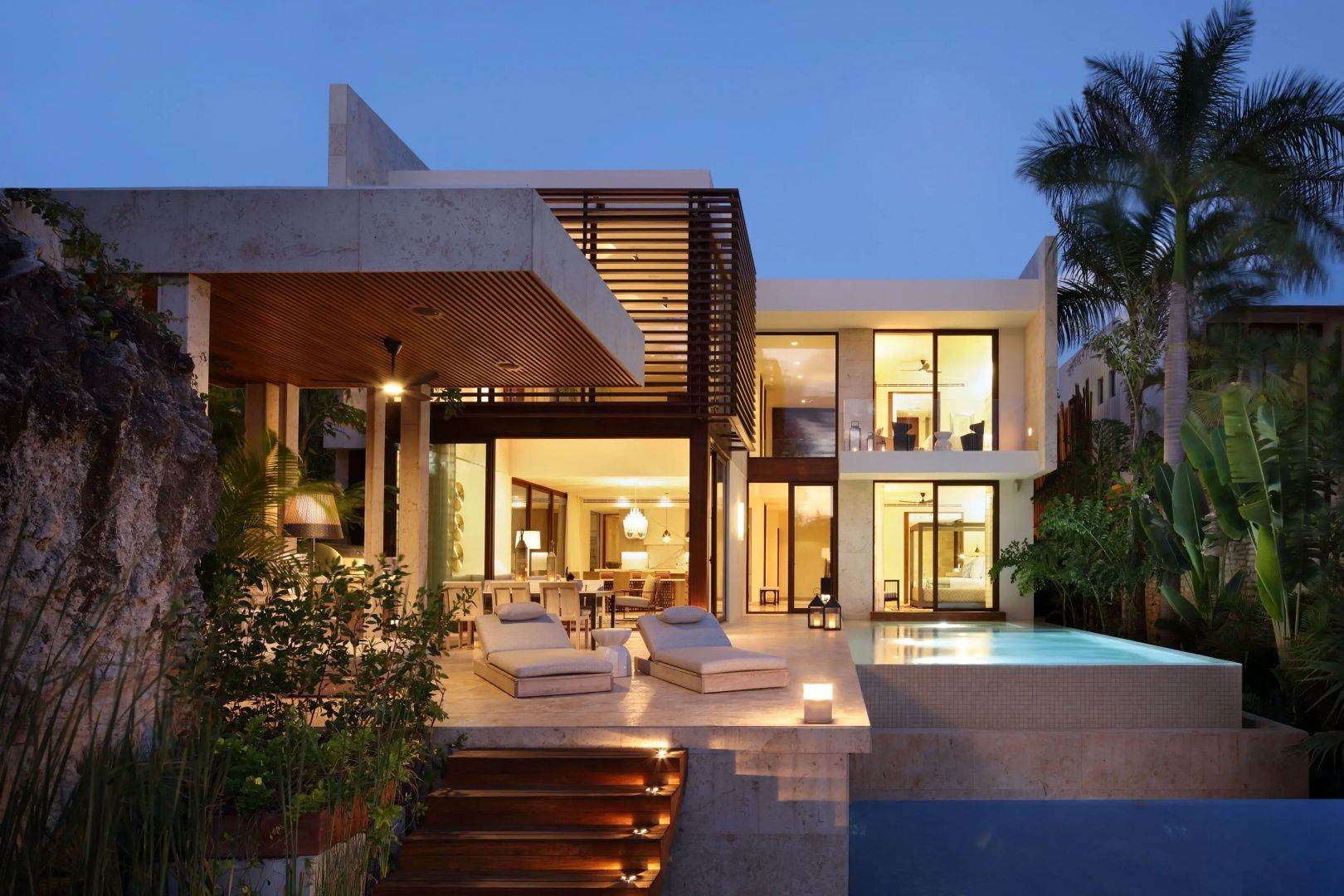 Zweistöckige moderne Villa bei Dämmerung mit beleuchteter Terrasse, Pool und Liegen.