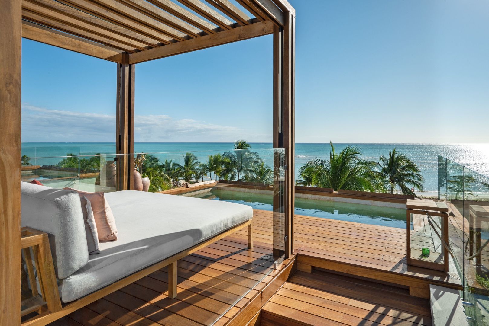 Sonnendeck aus Holz mit Tagesbett und kleinem Pool mit Blick auf Palmen und das Meer.