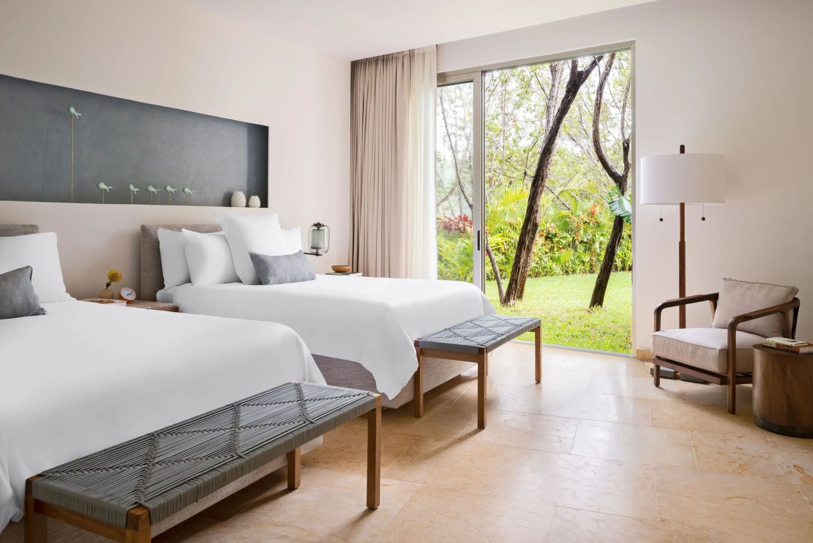 Schlafzimmer mit zwei Doppelbetten, Sitzbänken und Blick ins Grüne durch große Fenster.