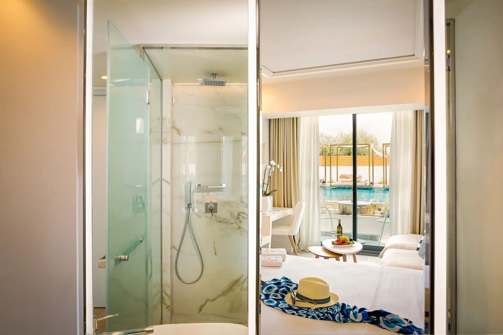 Einblick in das Hotelzimmer mit verglaster Dusche, Doppelbett und Poolblick.