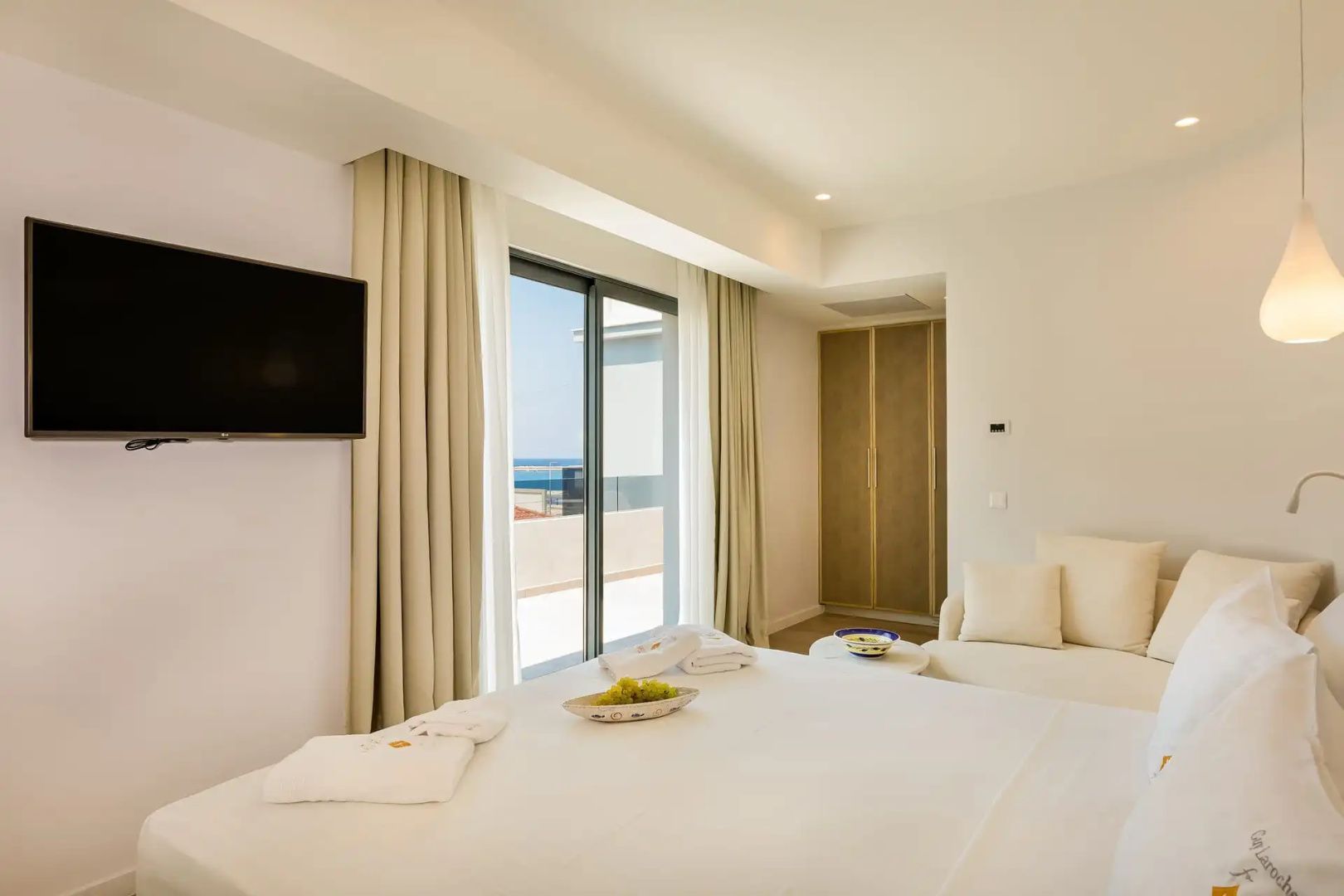 Schlafzimmer mit weiß gedecktem Bett, Flachbildfernseher und Schiebetür zum Balkon.