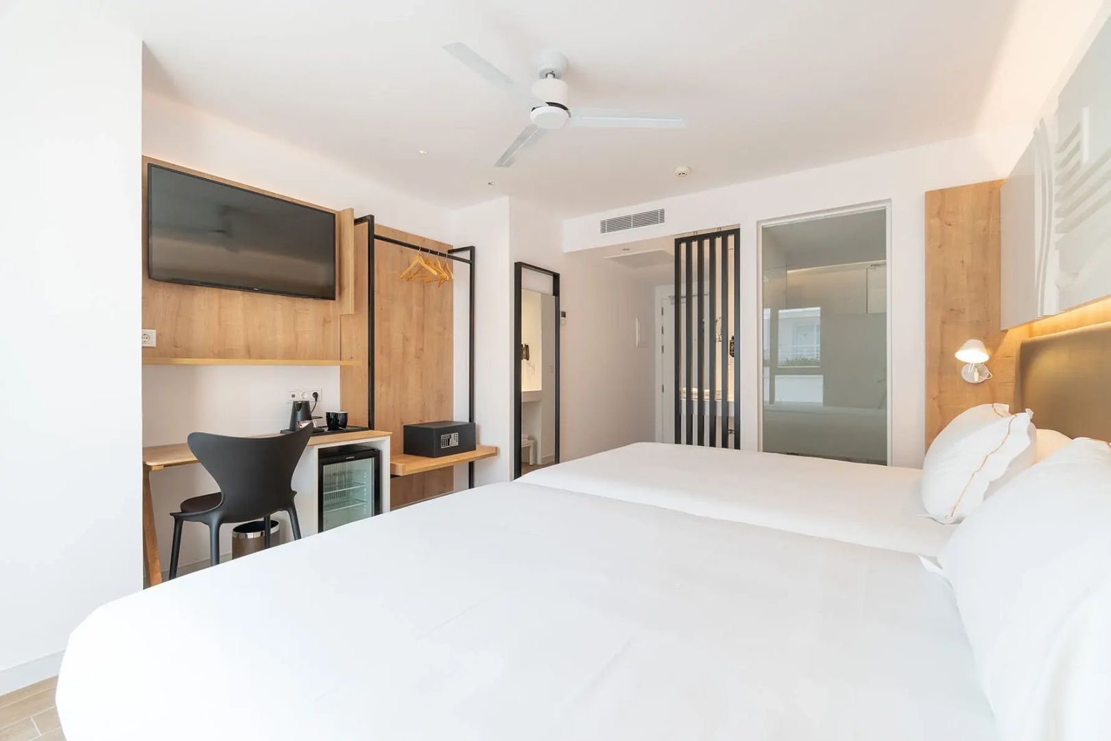 Helles Hotelzimmer mit Doppelbett, Fernseher an der Wand, Minibar und modernem Holzregal.