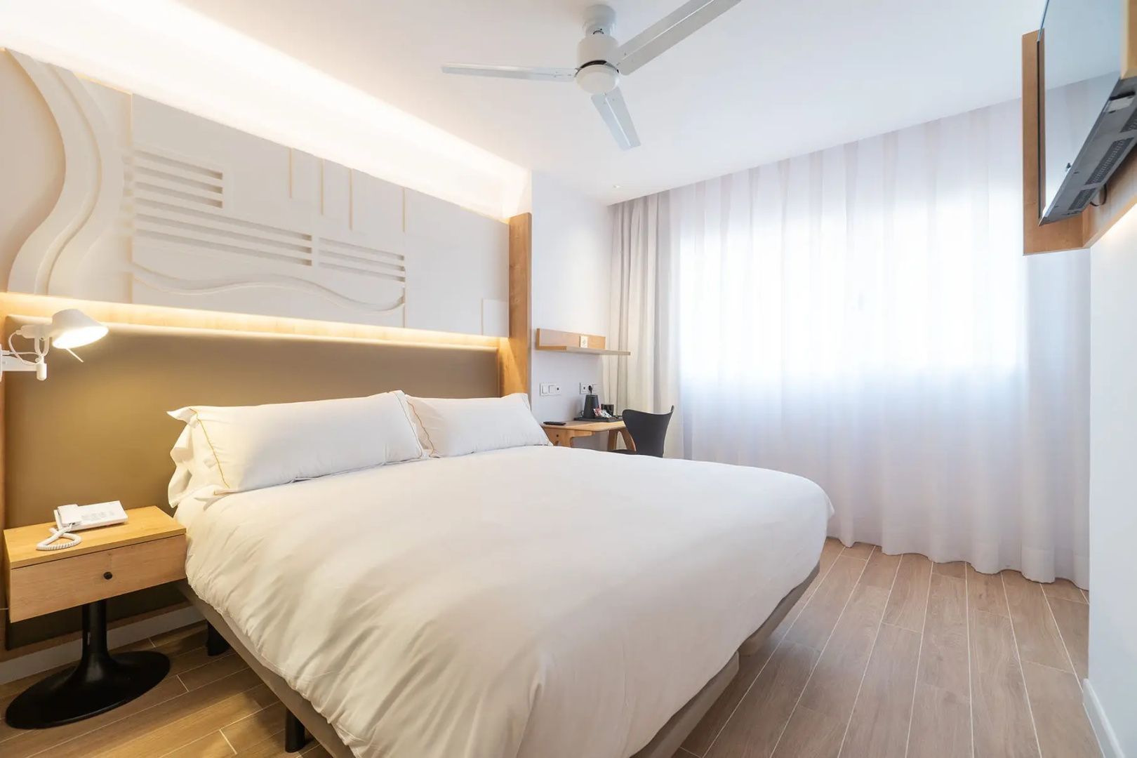 Hell eingerichtetes Doppelzimmer mit weißer Bettwäsche, Deckenventilator und Vorhängen.