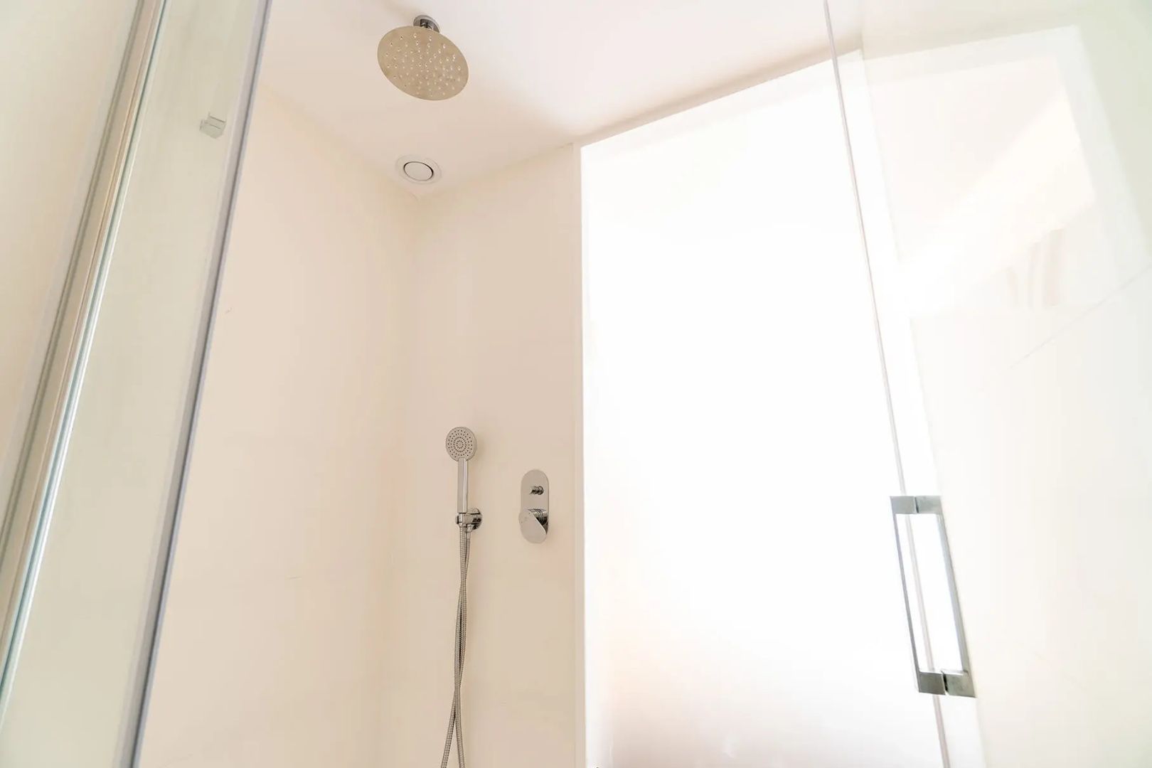 Hell geflieste Dusche im Badezimmer mit Regenduschkopf, Handbrause und Glaswand.