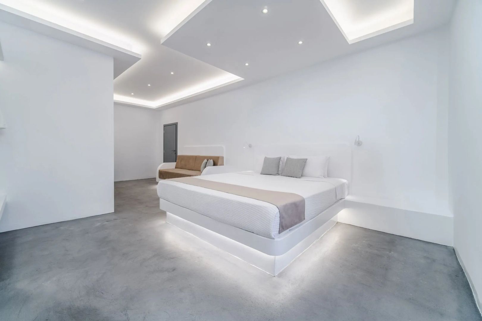 Weißes Hotelzimmer mit beleuchtetem Bett, integriertem Sofa und indirekter Deckenbeleuchtung.