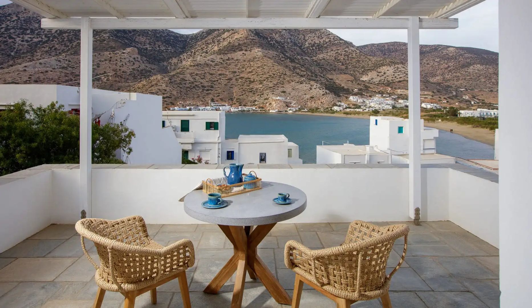 Überdachte Terrasse mit rundem Tisch, Stühlen und Blick auf eine Meeresbucht.