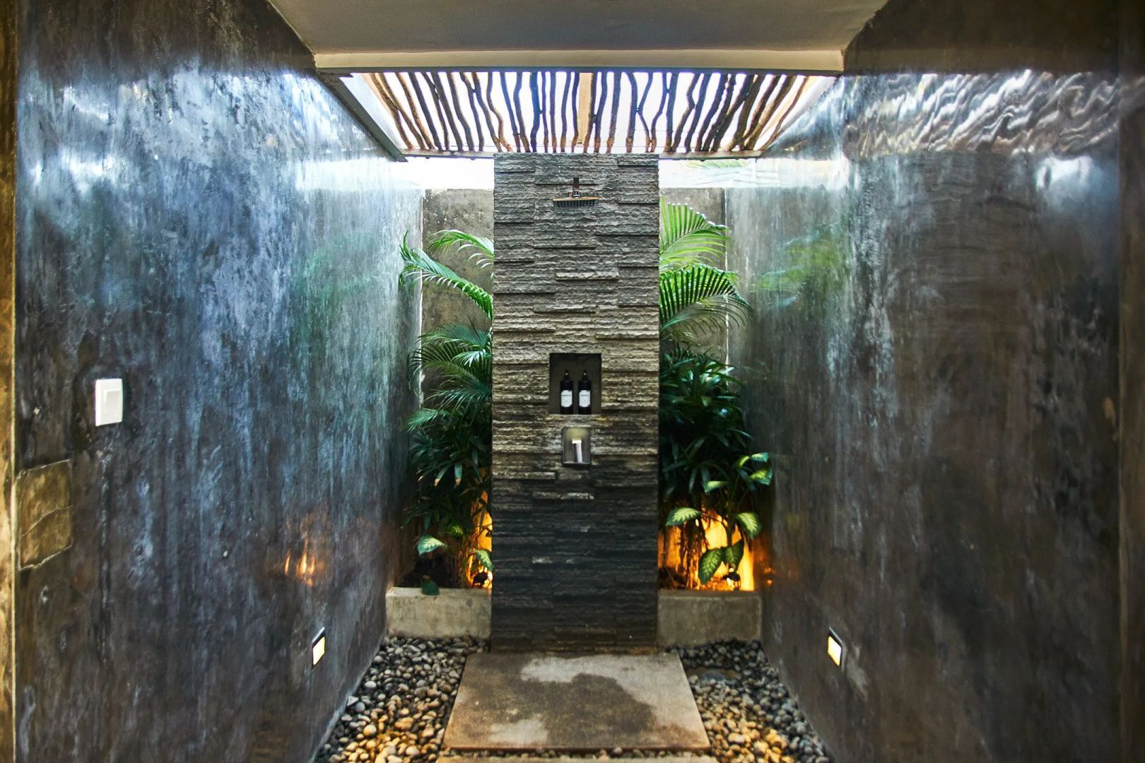 Regendusche im Außenbereich mit Natursteinwand, tropischen Pflanzen und Kieselsteinen.