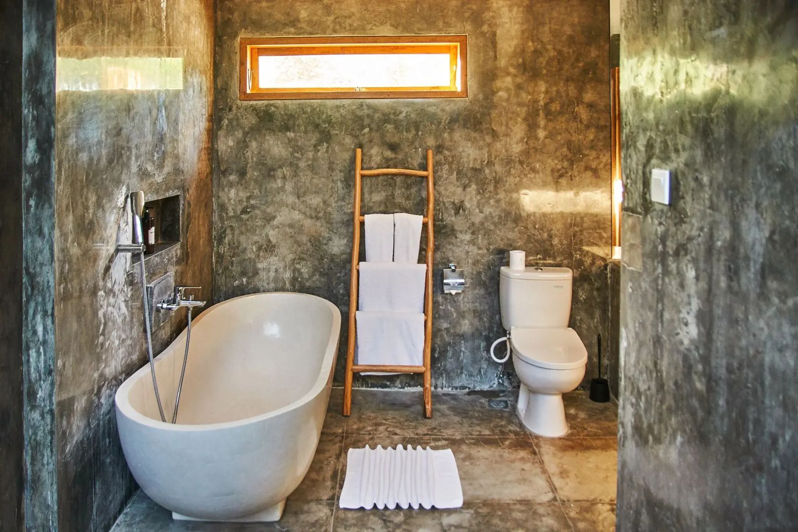 Badezimmer mit freistehender Badewanne, Toilette und Holzleiter an grauer Betonwand.
