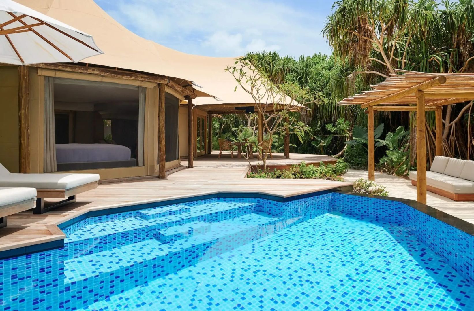 Privater Pool mit blauen Mosaikfliesen auf der Holzterrasse einer mit Reet gedeckten Villa.