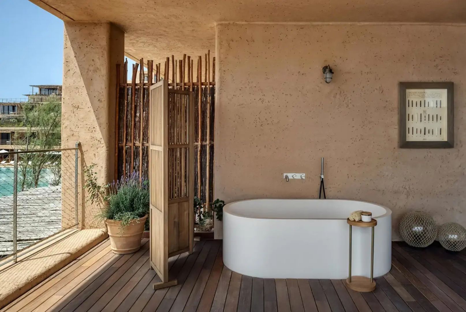 Freistehende weiße Badewanne auf einer Holzterrasse vor beigefarbener Wand mit Sichtschutz.