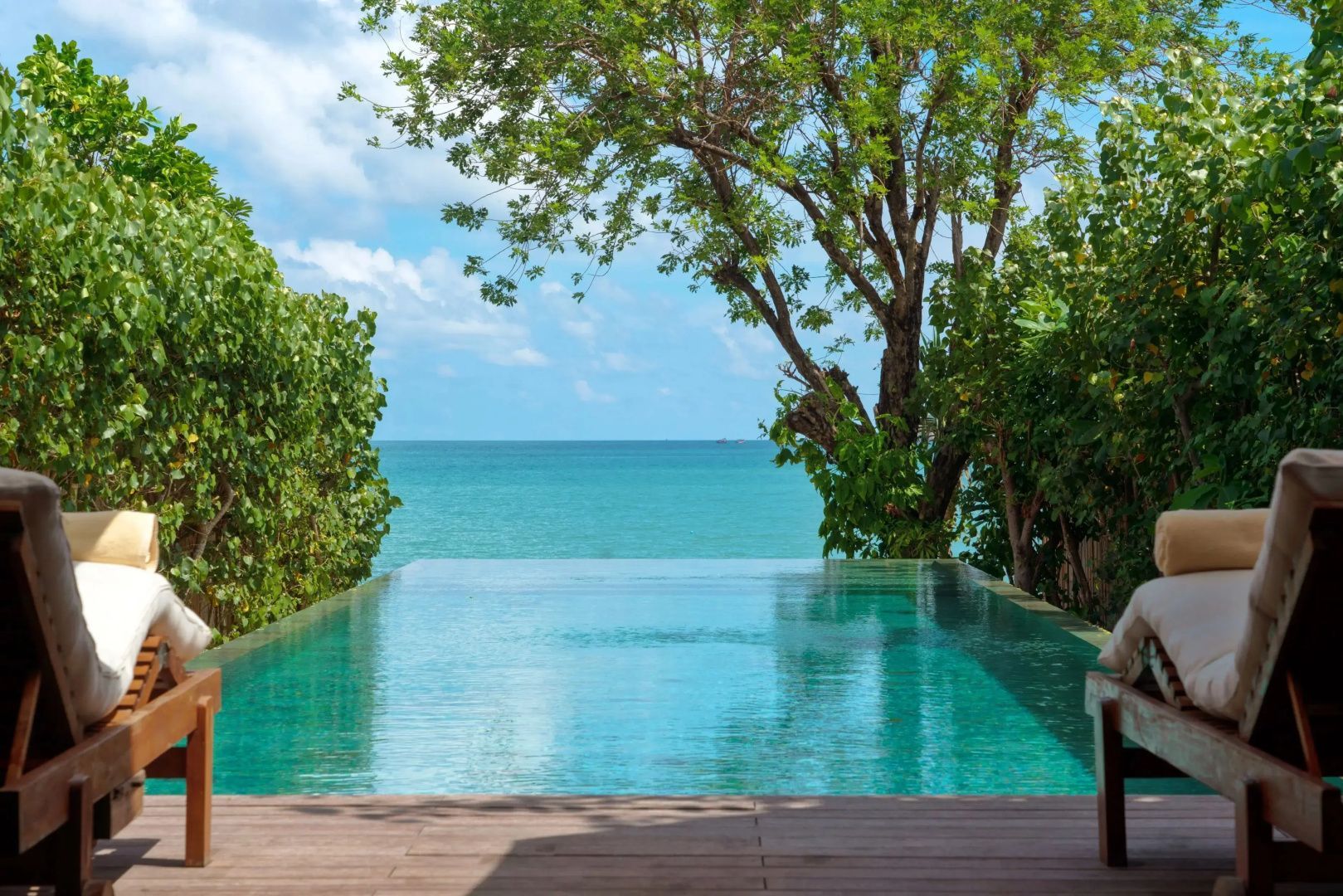 Blick über das Wasser eines Infinitypools auf das ruhige Meer und grüne Tropenpflanzen.