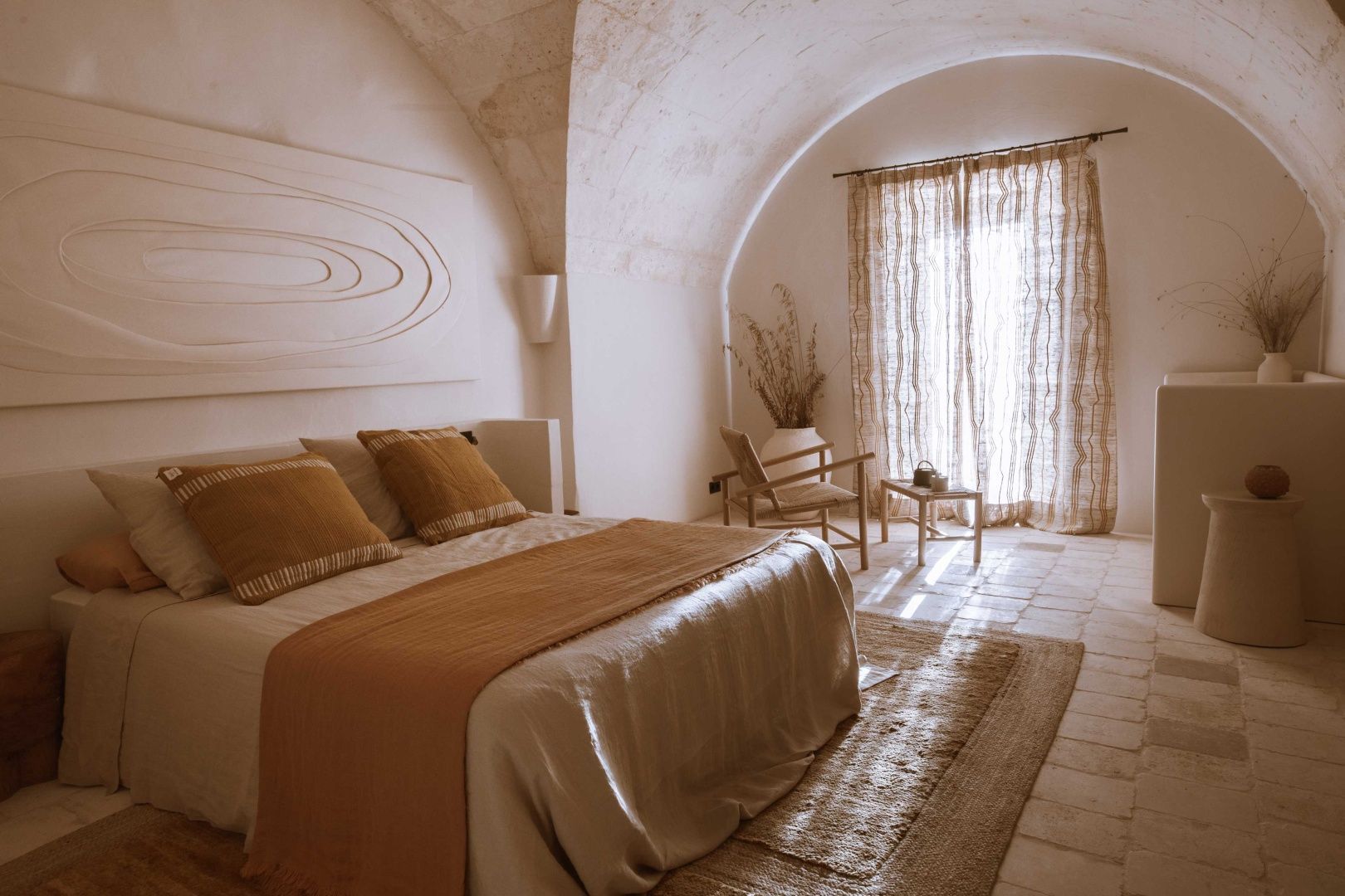 Schlafzimmer unter einem weißen Steingewölbe mit Doppelbett und Holzstuhl am Fenster.