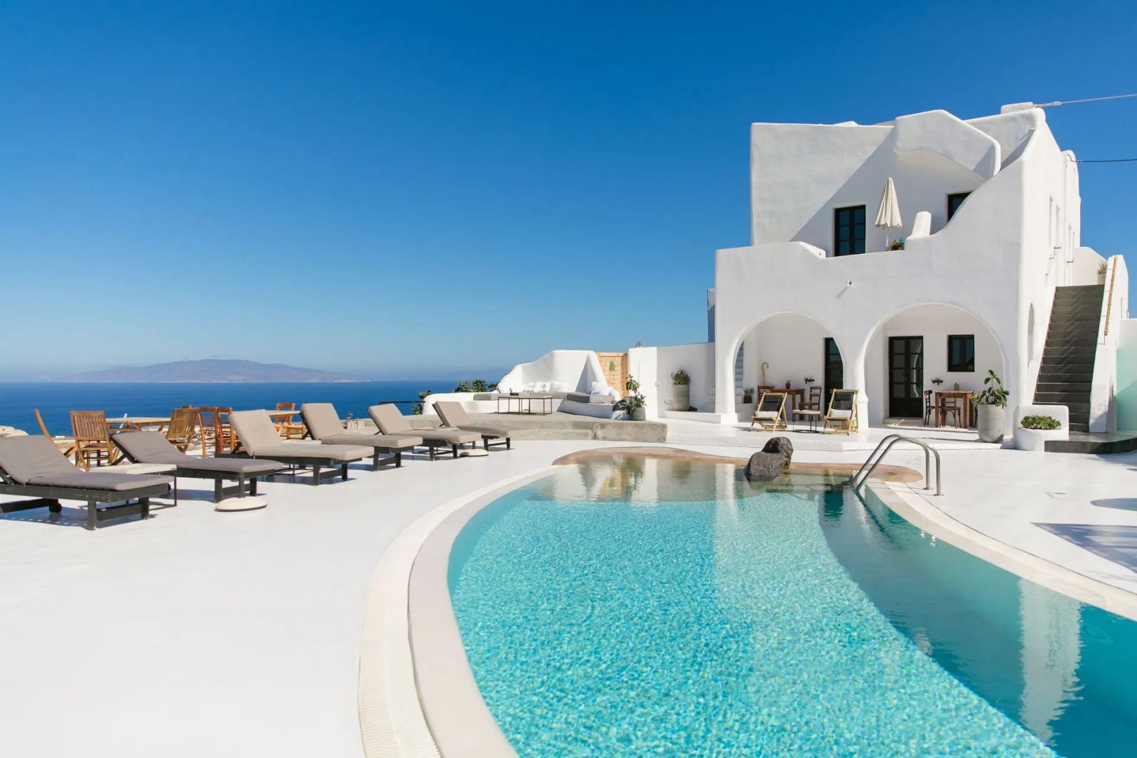 Außenpool mit Sonnenliegen auf weißer Terrasse und weitem Blick auf das Meer.