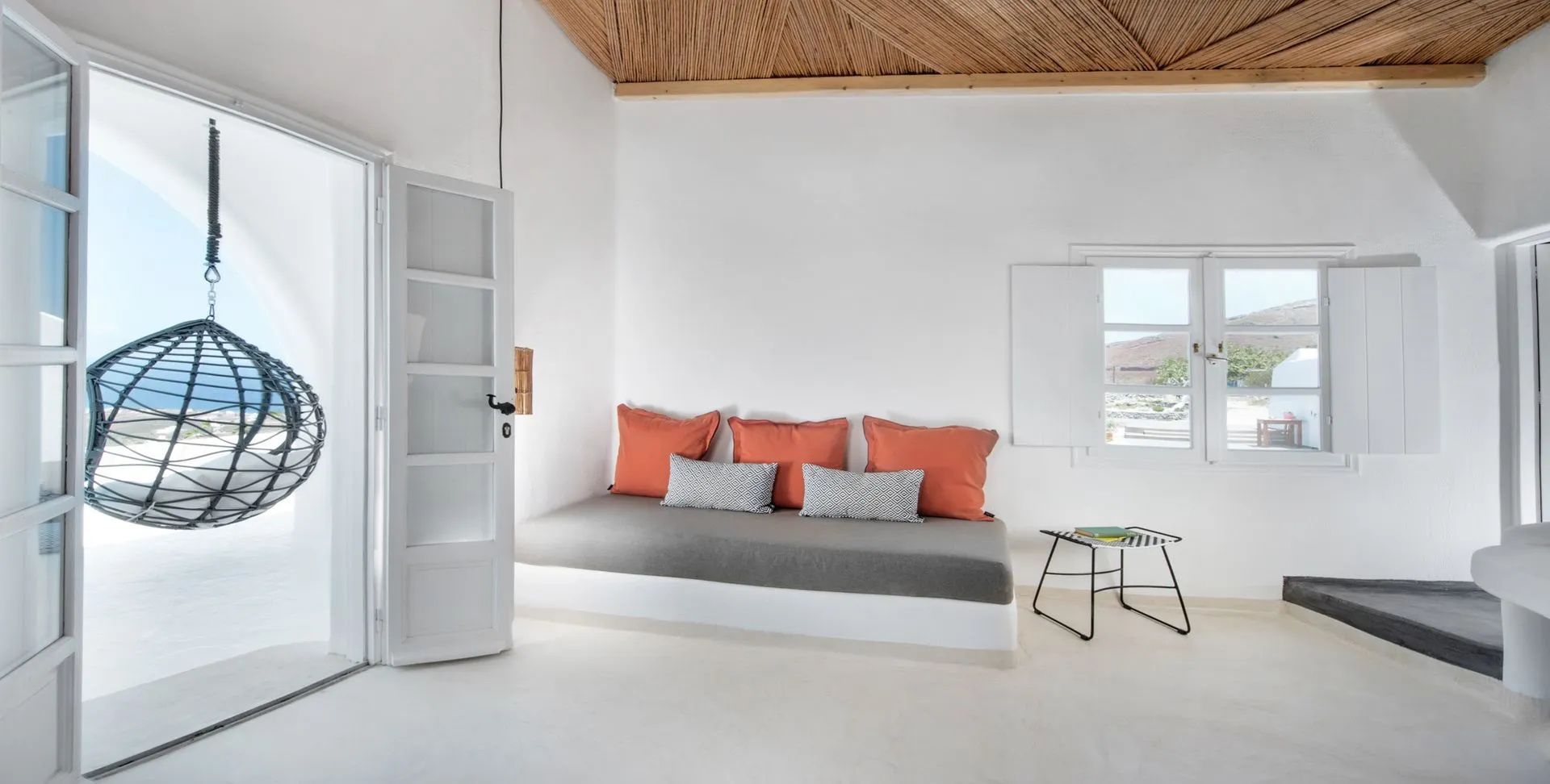 Ein Sitzbereich mit grauer Polsterbank, orangefarbenen Kissen und Holzdecke in weißem Raum.