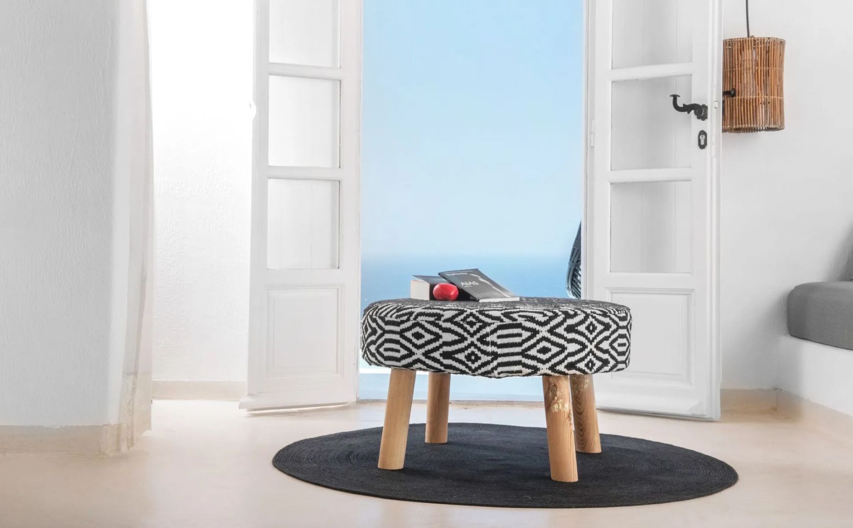 Gemusterter Hocker auf rundem Teppich vor geöffneter Tür mit Blick auf das Meer.