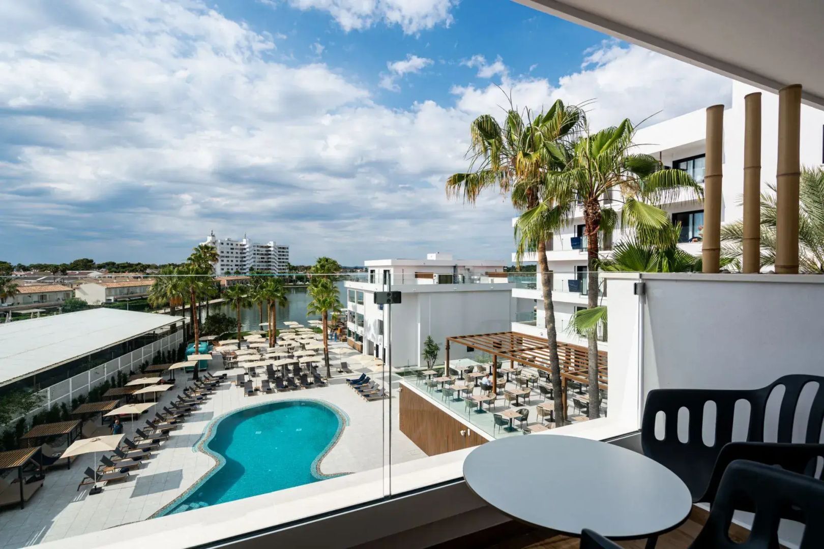 Blick vom Balkon mit Tisch auf die Poolanlage, Palmen und die umliegenden weißen Gebäude des Hotels.