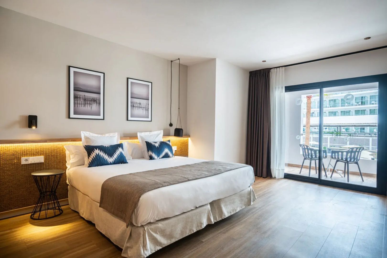 Doppelbett mit weißen Kissen und blauen Akzenten neben einer Balkontür in einem hellen Hotelzimmer.