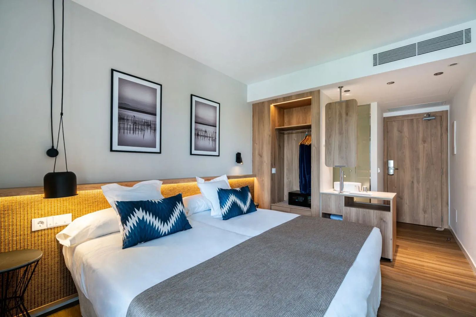 Helles Hotelzimmer mit Doppelbett, beigem Überwurf, Holzschrank und offenem Waschtisch.